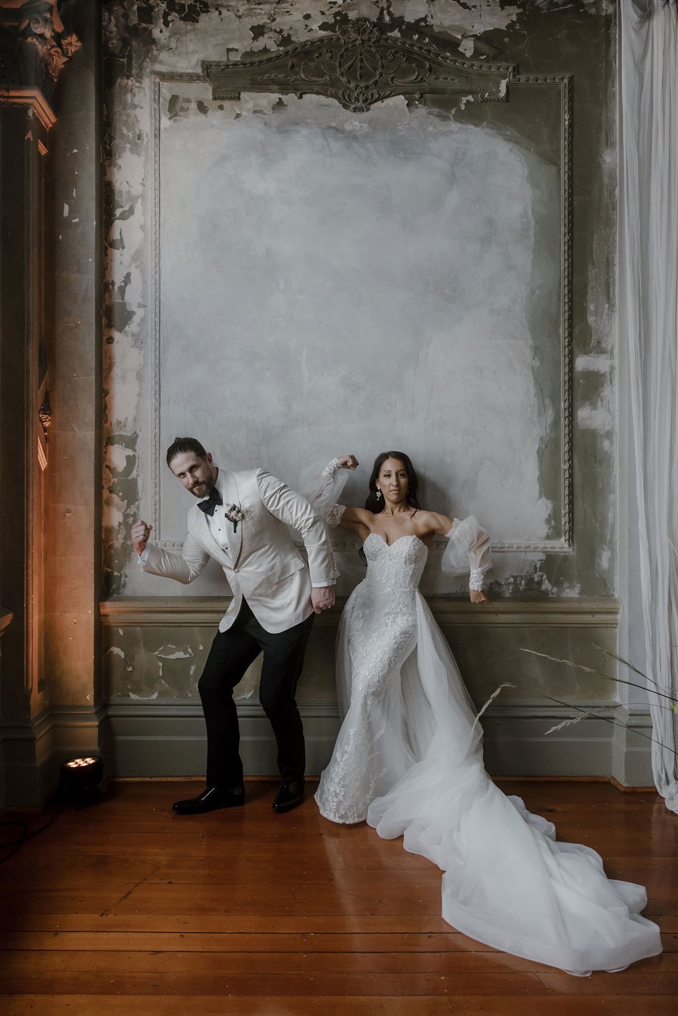 A playful and artistic wedding portrait of the bride and groom at their George Ballroom wedding in St Kilda. The groom, dressed in a cream tuxedo jacket with a black bow tie, strikes a fun, exaggerated muscle-flexing pose. The bride, wearing a stunning lace wedding gown with a long, flowing train and sheer sleeves, mirrors his pose, leaning against the vintage, weathered walls of the ballroom. The couple's lightheartedness contrasts with the grand, textured setting, capturing a unique and memorable moment at their stylish and elegant wedding.