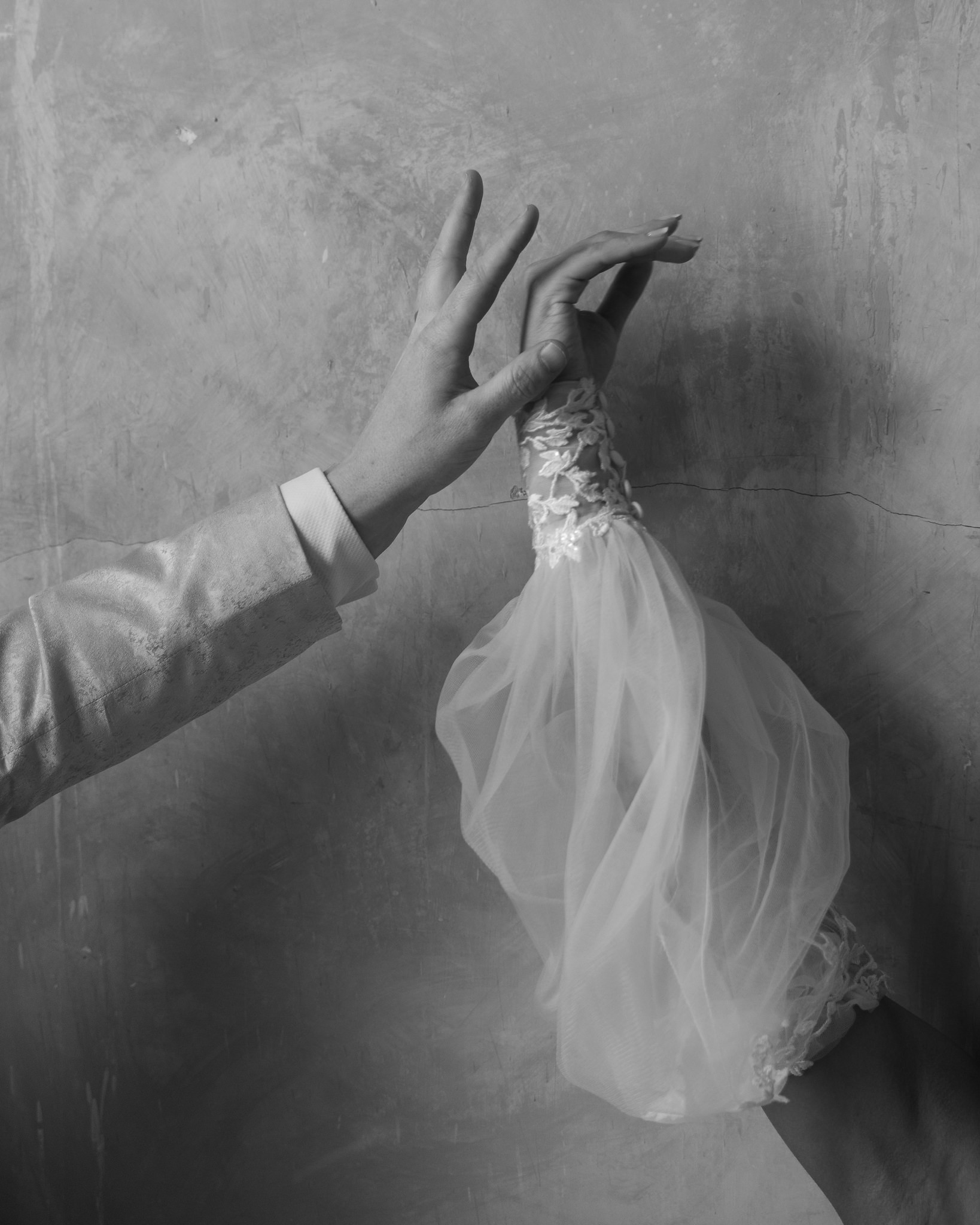A black and white close-up image of a bride and groom gently holding hands against a textured wall at their George Ballroom wedding in St Kilda. The groom’s hand, dressed in a light-colored tuxedo jacket, delicately reaches towards the bride’s hand, which is adorned in a sheer, lace-trimmed sleeve. The image captures an intimate, artistic moment of connection and tenderness, emphasizing the intricate details of the wedding attire and the romantic atmosphere at the historic George Ballroom in St Kilda.