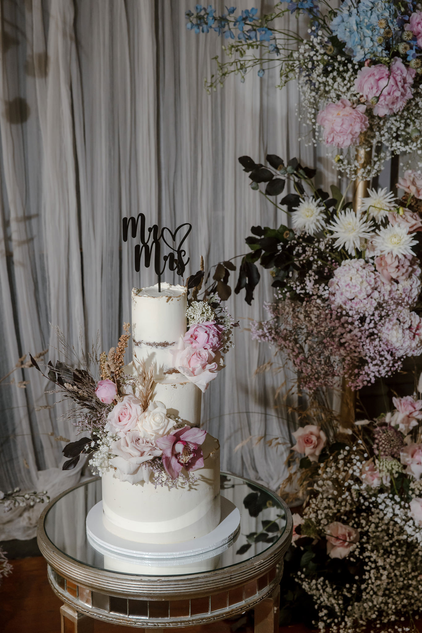 A beautifully decorated three-tiered wedding cake displayed at a George Ballroom wedding in St Kilda. The cake features a minimalist white design adorned with fresh floral accents, including soft pink and white roses, orchids, and dried foliage. A black 'Mr & Mrs' cake topper sits on the top tier, adding a personalized touch. The cake is placed on a round mirrored table, surrounded by lush floral arrangements of hydrangeas, roses, and greenery that complement the romantic and elegant atmosphere of the ballroom wedding.