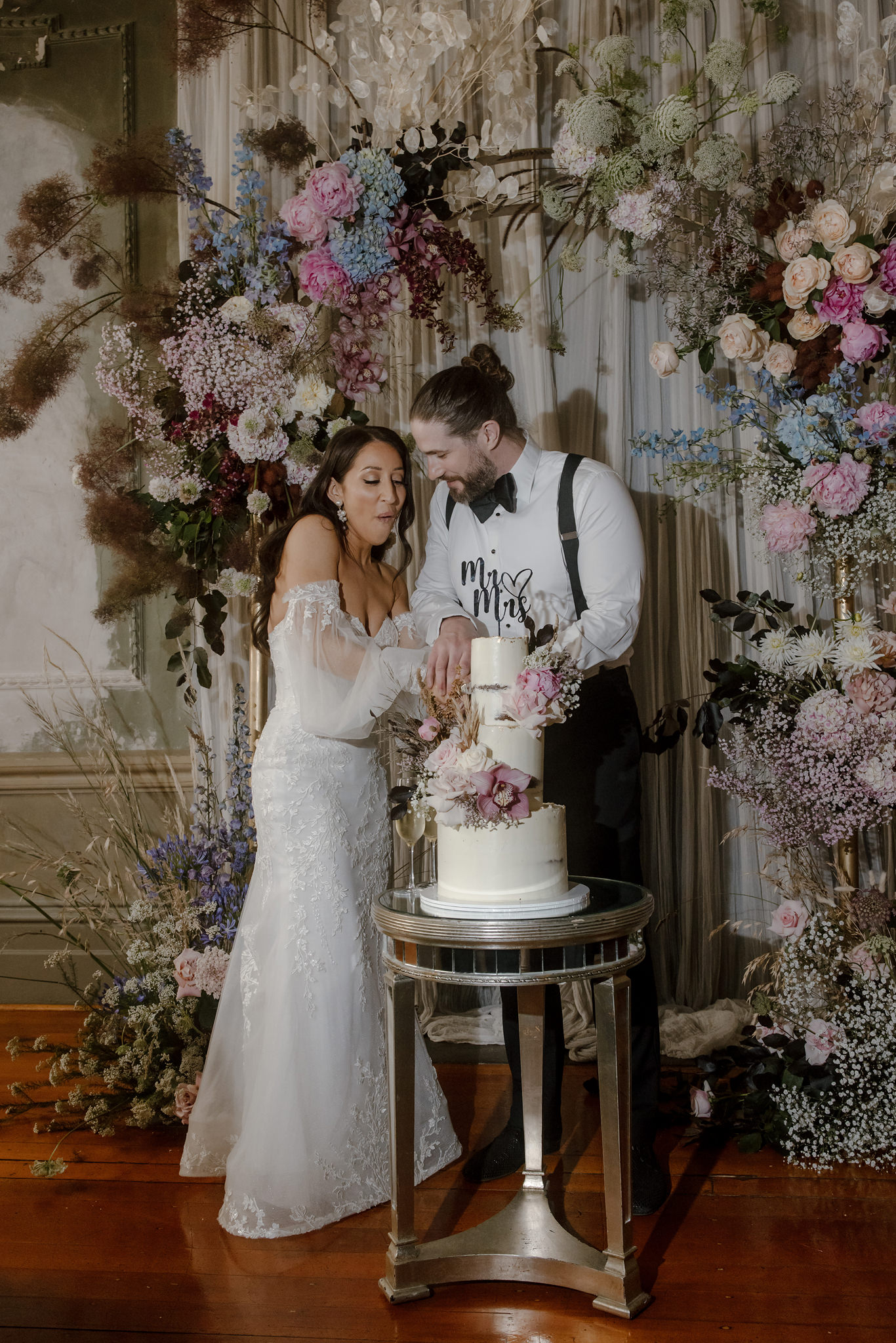 A joyful moment captured at a George Ballroom wedding in St Kilda as the bride and groom cut their wedding cake together. The bride, dressed in an elegant lace off-the-shoulder gown with sheer, voluminous sleeves, smiles with delight while helping the groom make the first cut. The groom, wearing suspenders and a white shirt with a black bow tie, beams as they share this special moment. The tiered cake, decorated with fresh flowers in shades of pink and cream, sits on a mirrored table, with a 'Mr & Mrs' topper adding a romantic touch. Behind them, a stunning floral display of hydrangeas, roses, and greenery complements the sophisticated ambiance of the George Ballroom wedding.