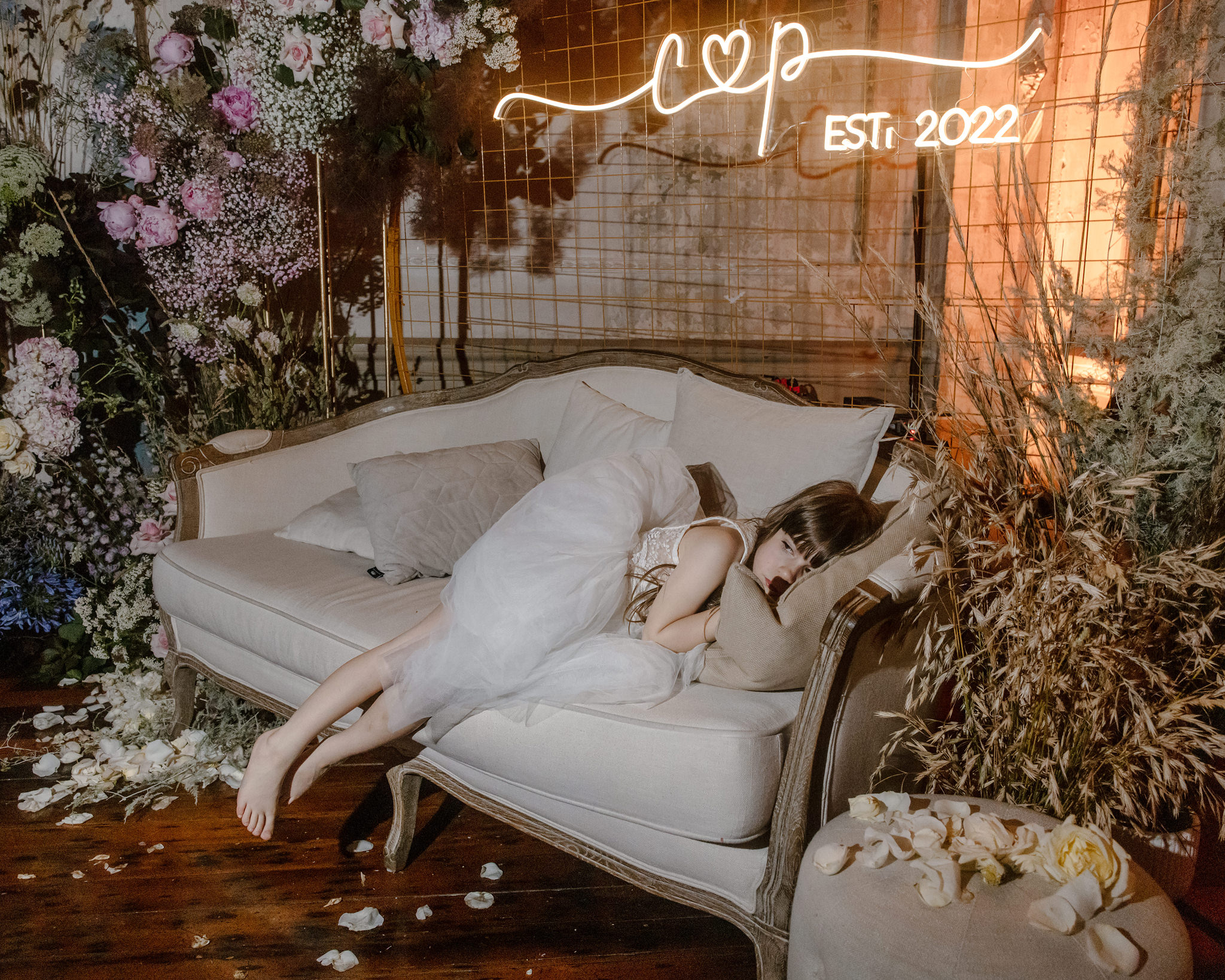 A candid moment at a George Ballroom wedding in St Kilda, featuring a young flower girl asleep on a vintage couch surrounded by an elaborate floral display. She lies peacefully with her legs hanging over the side of the cream-colored sofa, dressed in a delicate white dress. Behind her, a custom neon sign reads 'C&P Est. 2022,' and the space is decorated with lush arrangements of roses, baby’s breath, and dried florals in soft tones of pink, purple, and white. Rose petals are scattered across the floor, adding to the whimsical and serene atmosphere of the wedding reception at the historic George Ballroom in St Kilda.