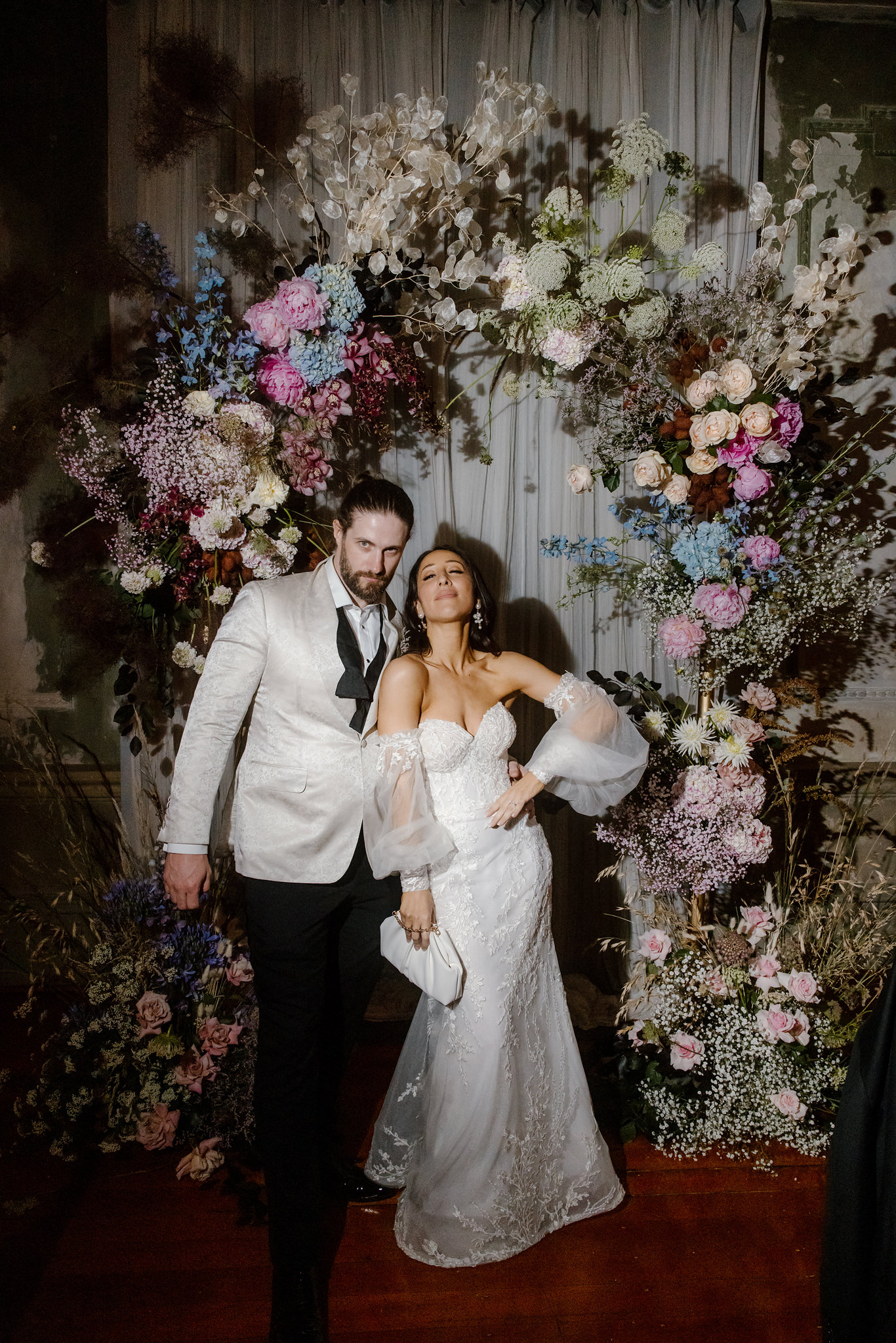 A bride and groom stand confidently in front of a lush and dramatic floral arch at their wedding in the George Ballroom, St Kilda. The groom wears a stylish cream tuxedo jacket paired with a black bow tie, his hair slicked back in a modern yet classic look, while the bride stuns in an elegant, off-the-shoulder lace wedding gown with voluminous sheer sleeves, holding a chic white clutch. The couple exudes playful confidence as they strike a bold pose together, framed by an exquisite backdrop of cascading florals featuring hydrangeas, roses, baby’s breath, and other lush greenery in a mix of soft pinks, blues, and earthy tones. The moody, romantic ambiance of the George Ballroom enhances the overall vibe of sophistication and modern elegance, capturing a distinctively stylish wedding moment in St Kilda.