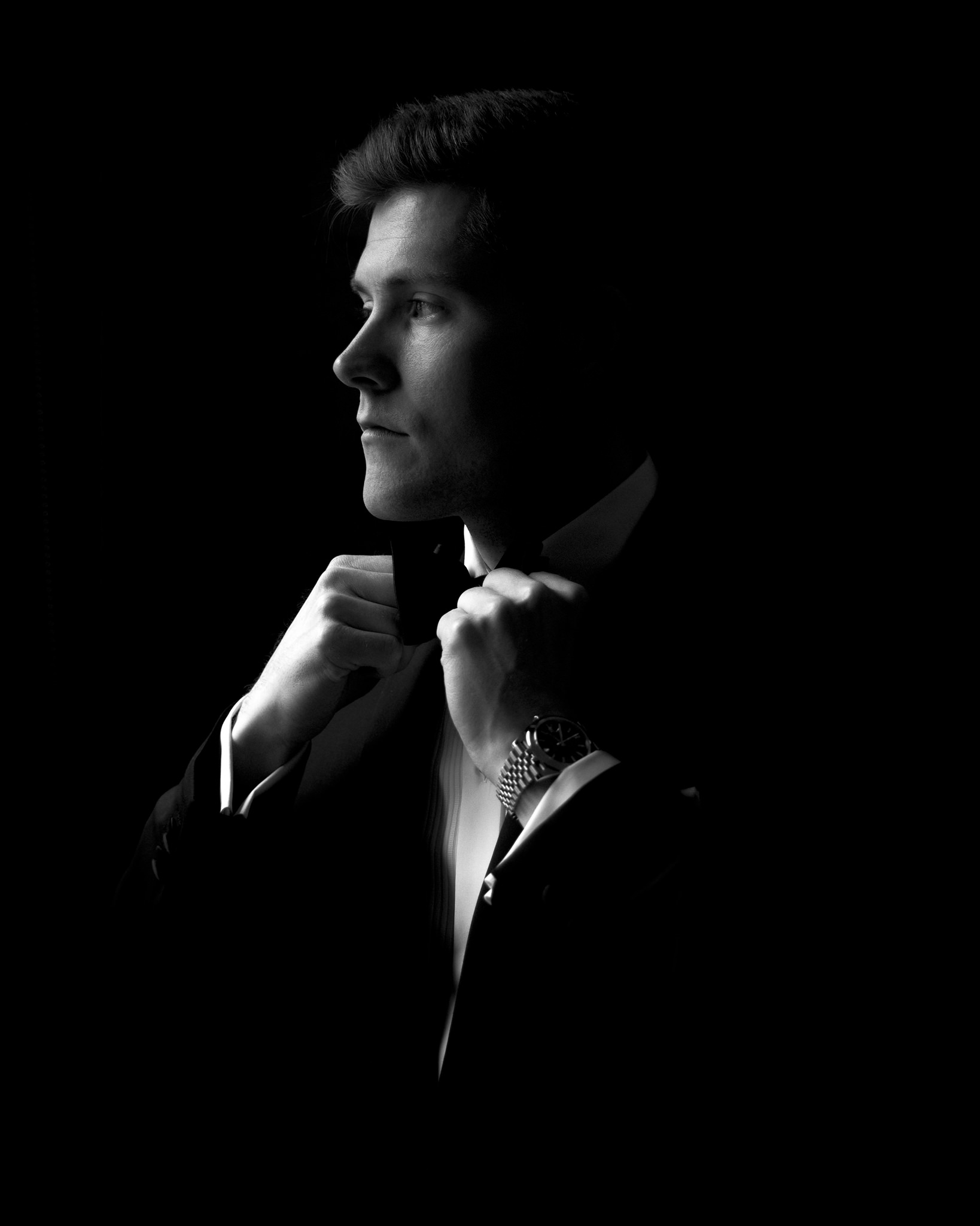 The image is a dramatic black and white portrait of a young man in formal attire, focusing on his profile from the chest upwards. He is in the process of adjusting his bow tie with both hands, exuding an air of sophistication and elegance. The lighting is meticulously crafted to highlight the contours of his face and the details of his tuxedo, while the background remains shrouded in darkness, enhancing the intensity and mood of the moment. His expression is calm and contemplative, suggesting a moment of preparation or reflection. The watch on his wrist adds a touch of luxury and practicality, visible due to the striking contrast in lighting. This portrait captures a personal and intimate moment, typical of wedding day preparations.