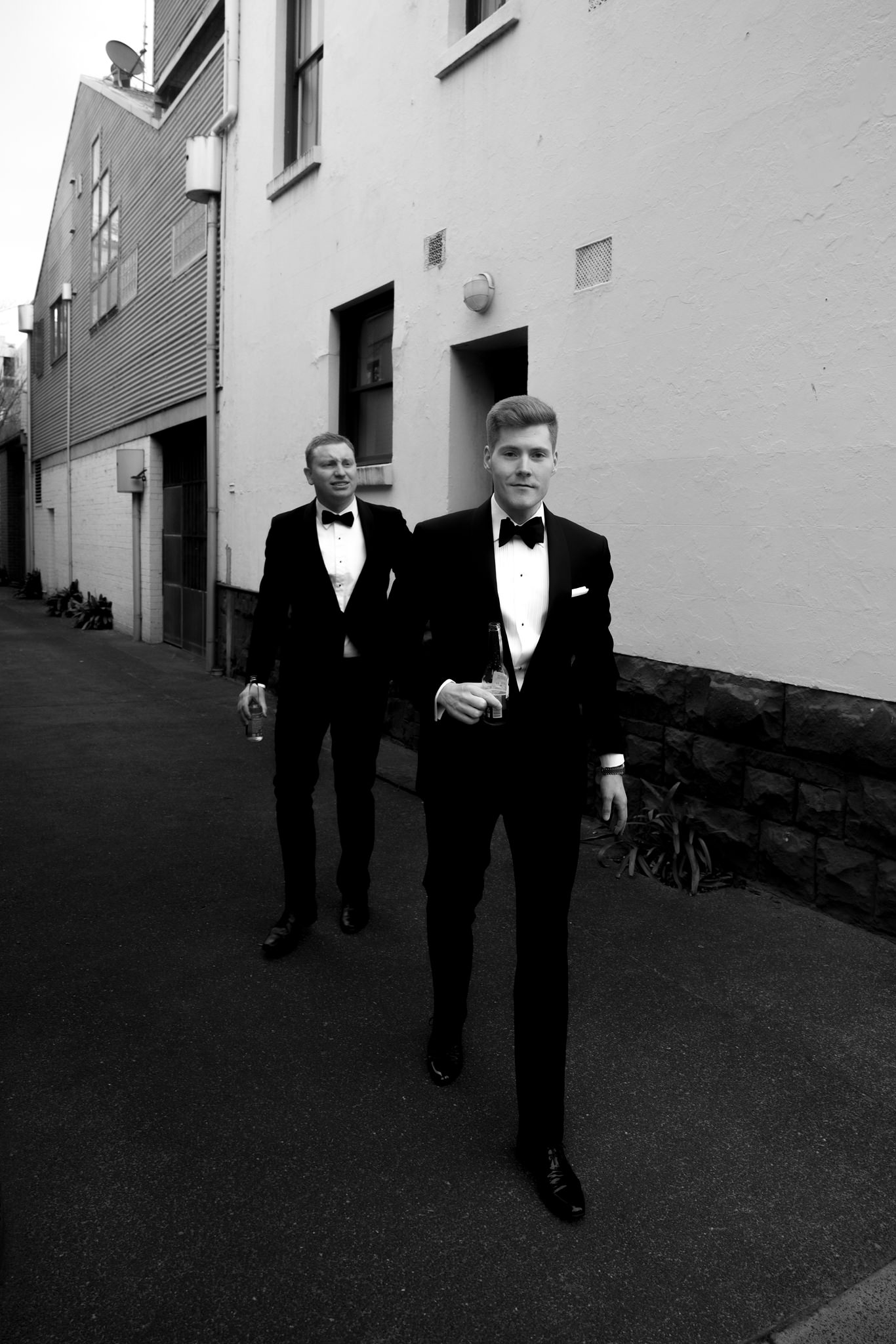 The image is a stylish black and white photograph featuring two men dressed in elegant black tuxedos with bow ties, walking along an urban alleyway. The man in the foreground is holding a glass of champagne and directly facing the camera with a confident, relaxed expression. The background features a mix of residential and industrial building facades, providing a contrasting, textured environment that adds an edgy, modern feel to the traditional wedding attire. The lighting is soft, illuminating the subjects while casting gentle shadows, which accentuates the details of their suits and the casual yet refined atmosphere of the moment. This photograph captures a candid, possibly celebratory moment during a wedding event, blending classic style with an unconventional setting.