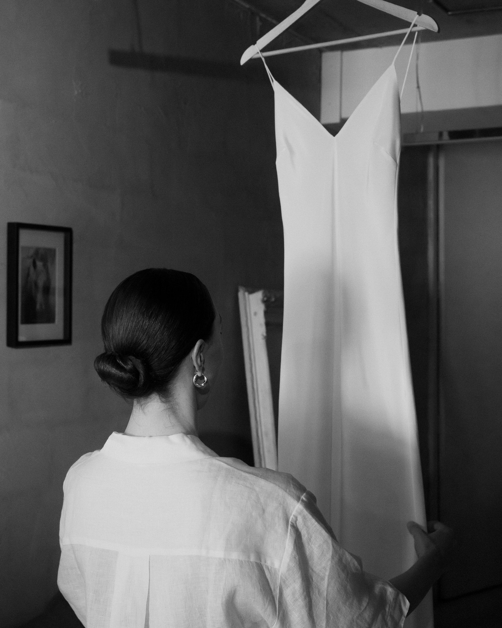 The image is a striking black and white photograph capturing a bride as she contemplates her wedding dress. The dress, a simple yet elegant white gown with a deep V-neckline, hangs on a wooden hanger from a ceiling-mounted rail, set against a rough, textured wall. The bride, viewed from the back, is dressed in a white linen shirt, and her hair is styled in a sleek bun. Her posture and the gentle placement of her hand on the dress convey a moment of reflection and anticipation. The environment suggests an urban loft or studio setting, with subdued lighting that focuses attention on the interaction between the bride and her dress. This image beautifully captures the intimate and personal moments of preparation before a wedding.