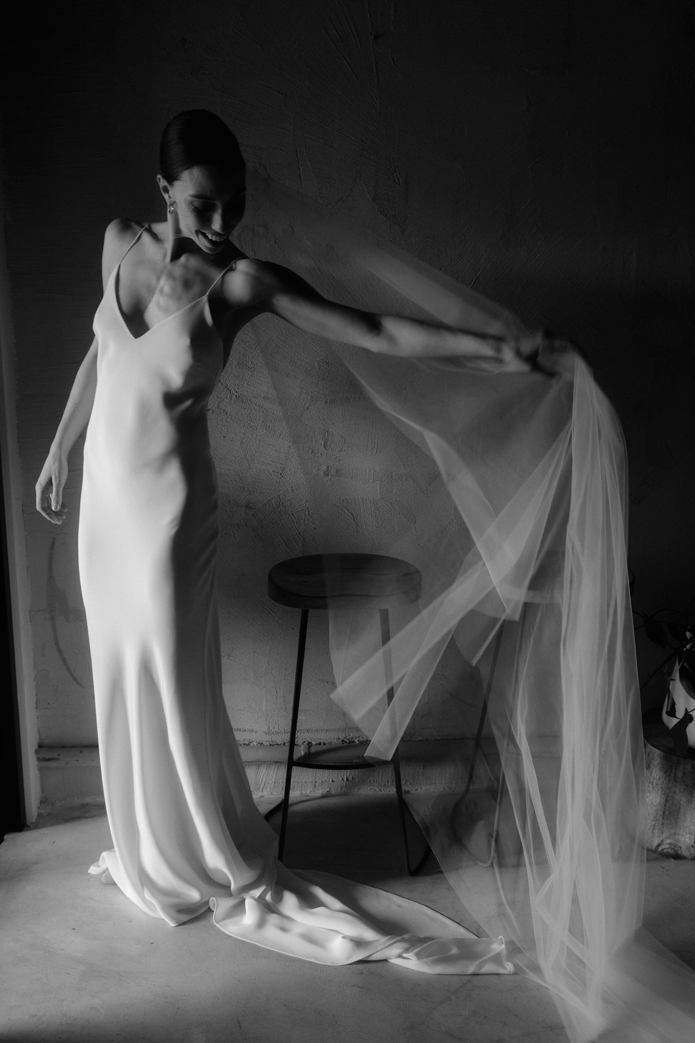 The image is a captivating black and white photograph of a bride playfully interacting with her wedding veil. She is standing in a softly lit room, clad in an elegant, form-fitting sleeveless wedding gown with a long flowing train. The bride's pose is dynamic and full of movement; she is smiling and looking down while gently pulling her veil to create an airy, billowing effect. The texture of the gown and the sheer fabric of the veil are beautifully contrasted against the rough, plastered wall behind her. Her hair is styled in a sleek bun, complementing her graceful demeanor. This photograph captures a joyful and light-hearted moment, highlighting the bride's excitement and the ethereal quality of her wedding attire.