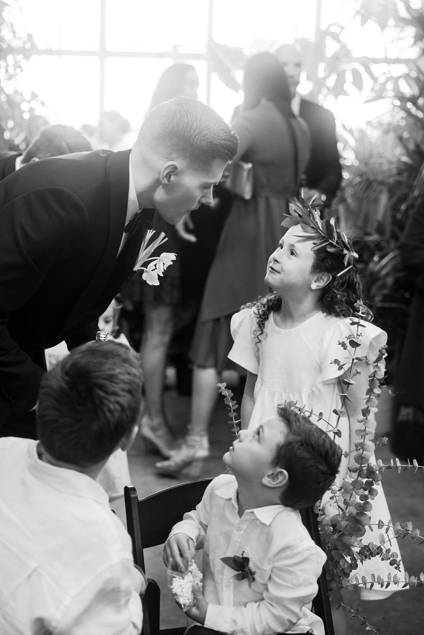 The image is a touching black and white photograph capturing a heartwarming moment between a groom and two young children at a wedding. The groom, dressed in a sharp black suit, is bending forward to interact with a little girl and a boy seated on a chair. The girl, adorned with a floral wreath in her hair, gazes up at the groom with a look of admiration and joy. She wears a white dress embellished with floral decorations that complement her wreath. The young boy, also in a white shirt and captivated by the interaction, looks up at the groom with a curious expression. This candid scene takes place in a lively setting, likely a reception area, filled with guests and natural light streaming through large windows, adding a dynamic and joyful atmosphere to the celebration. The photograph beautifully captures the genuine emotions and connections that often define the spirit of a wedding day.