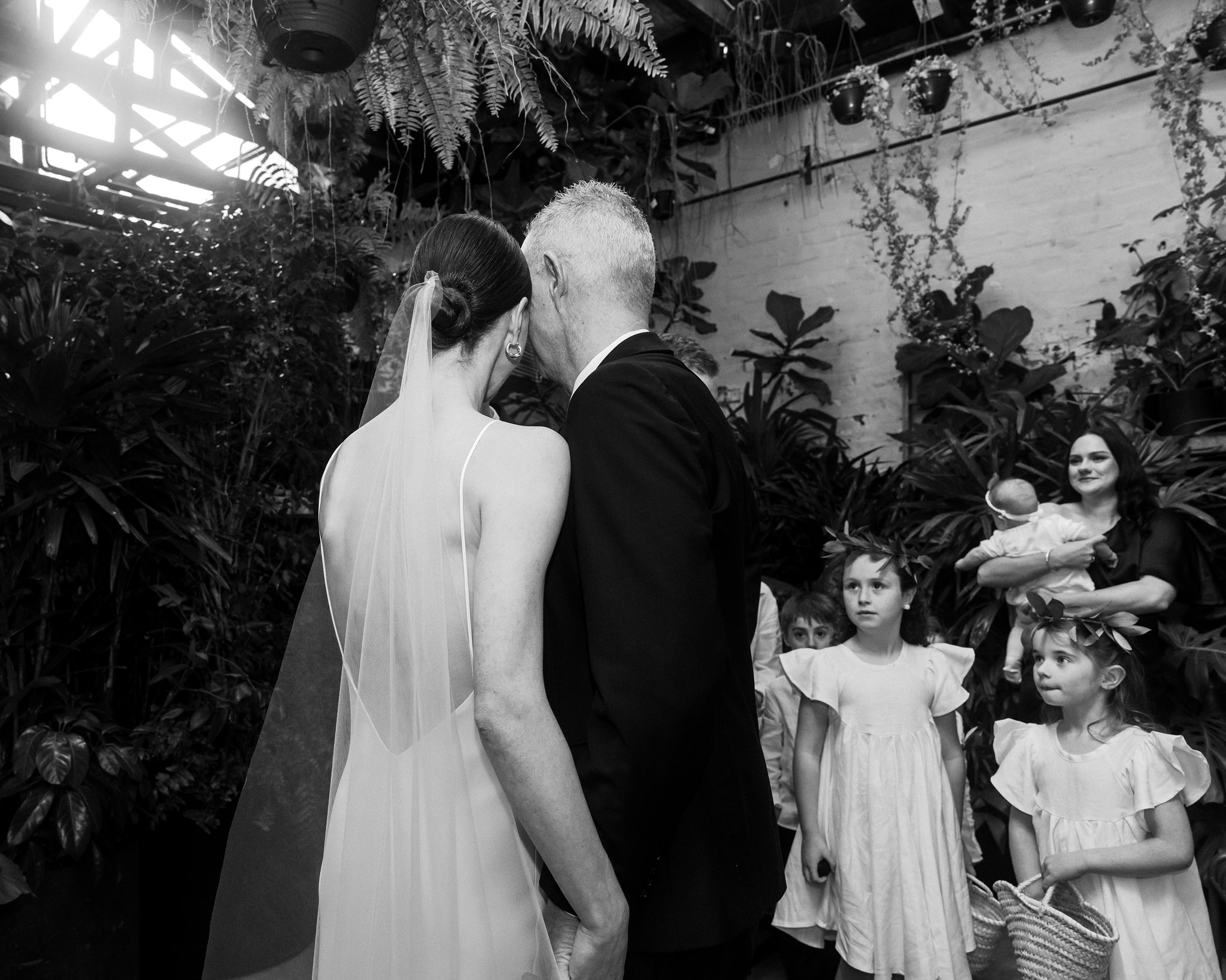 The image is a black and white photograph capturing a poignant moment at a wedding, featuring the bride and her father walking down the aisle, surrounded by an audience of guests in a lush, garden-like venue. The bride, dressed in a simple yet elegant sleeveless wedding gown with a delicate veil cascading down her back, is seen from behind as she walks closely with her father, who is dressed in a formal black suit. The setting is richly decorated with hanging plants, ferns, and a rustic brick wall, creating a verdant and intimate atmosphere. Several guests, including young children dressed in light, flowery dresses and floral headbands, watch the procession with varied expressions of awe and emotion. A woman holding a baby stands to the right, further emphasizing the family-oriented nature of the event. The overhead trellis partially visible at the top casts soft patterns of light and shadow across the scene, enhancing the emotional and serene quality of the moment.
