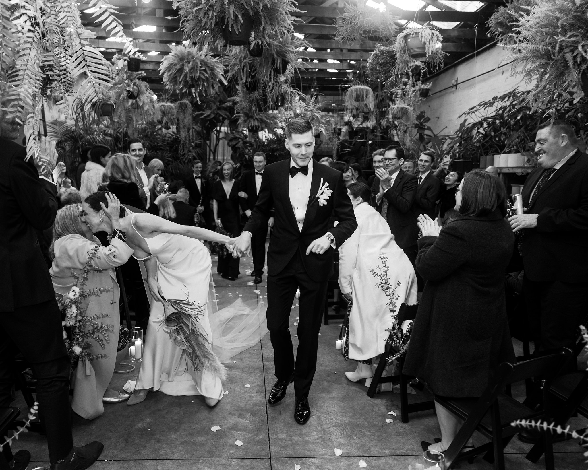 The image is a dynamic black and white photograph from a wedding reception, capturing the moment the bride and groom make their grand entrance, welcomed by a lively and celebratory crowd. The groom, dressed in an elegant black tuxedo, strides confidently forward with a broad smile, leading the way amidst applause from the guests. The bride, in a stylish white gown with floral embroidery, leans back joyfully to embrace an older woman, likely a family member, on her right. Guests are captured in various states of joy and applause, surrounding the couple with energy and happiness. The lush indoor garden setting, filled with hanging ferns and plants, adds a vibrant and lively backdrop to this celebratory moment. This photograph effectively conveys the joyous atmosphere of the wedding, with guests engaged and the couple at the heart of the celebration.