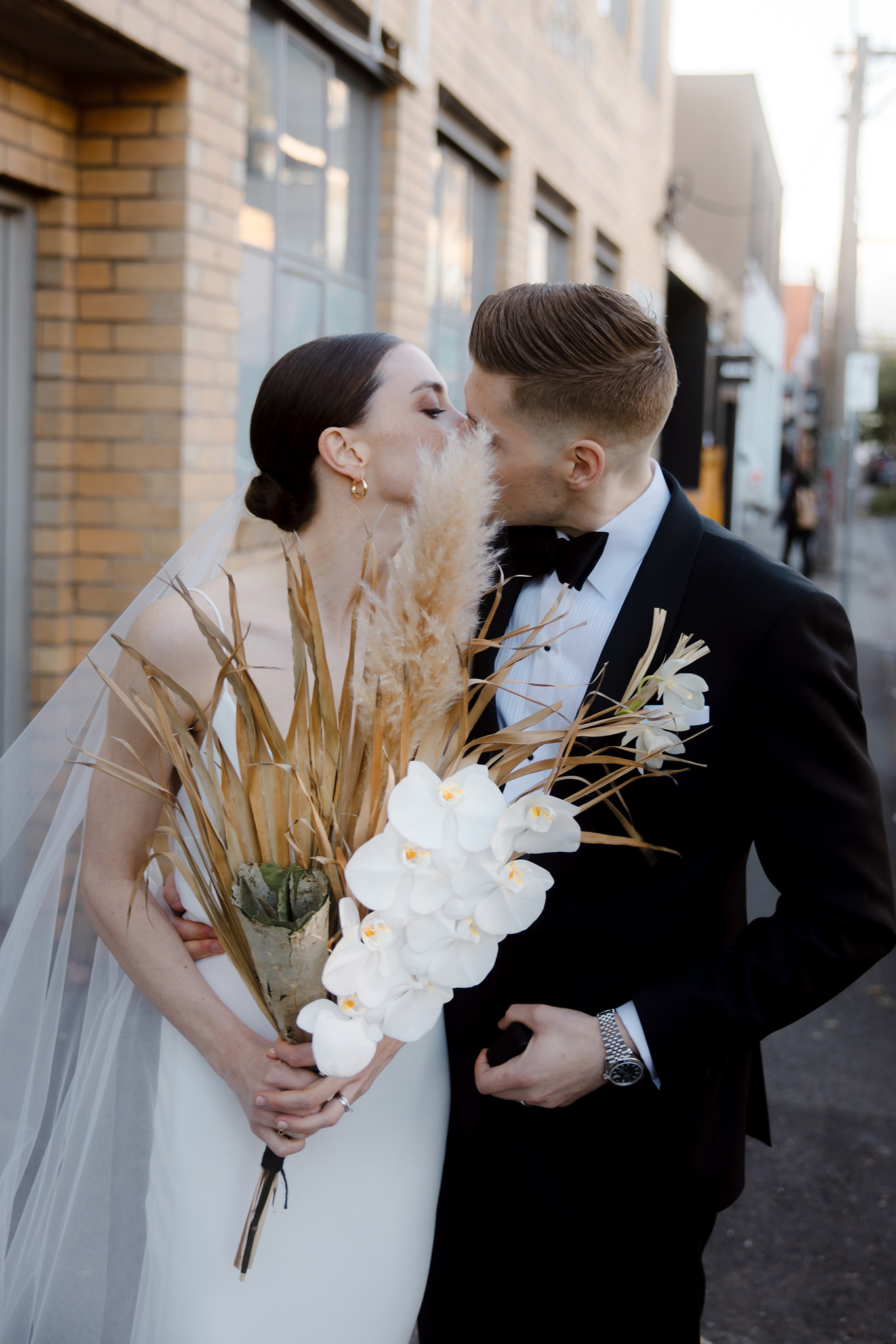 The image captures a tender moment between a bride and groom as they share a kiss on a city street. The bride, in a sleek, modern wedding dress, holds a unique bouquet featuring white orchids and dried pampas grass. The groom, dressed in a classic black tuxedo, gently kisses her cheek. The background shows a muted urban setting with a brick building and a narrow alley, adding a contemporary feel to the romantic scene. The couple's close embrace and the groom's hand gently touching the bride’s waist highlight their intimate connection. This photograph combines urban elegance with personal romance, reflecting a modern wedding style.