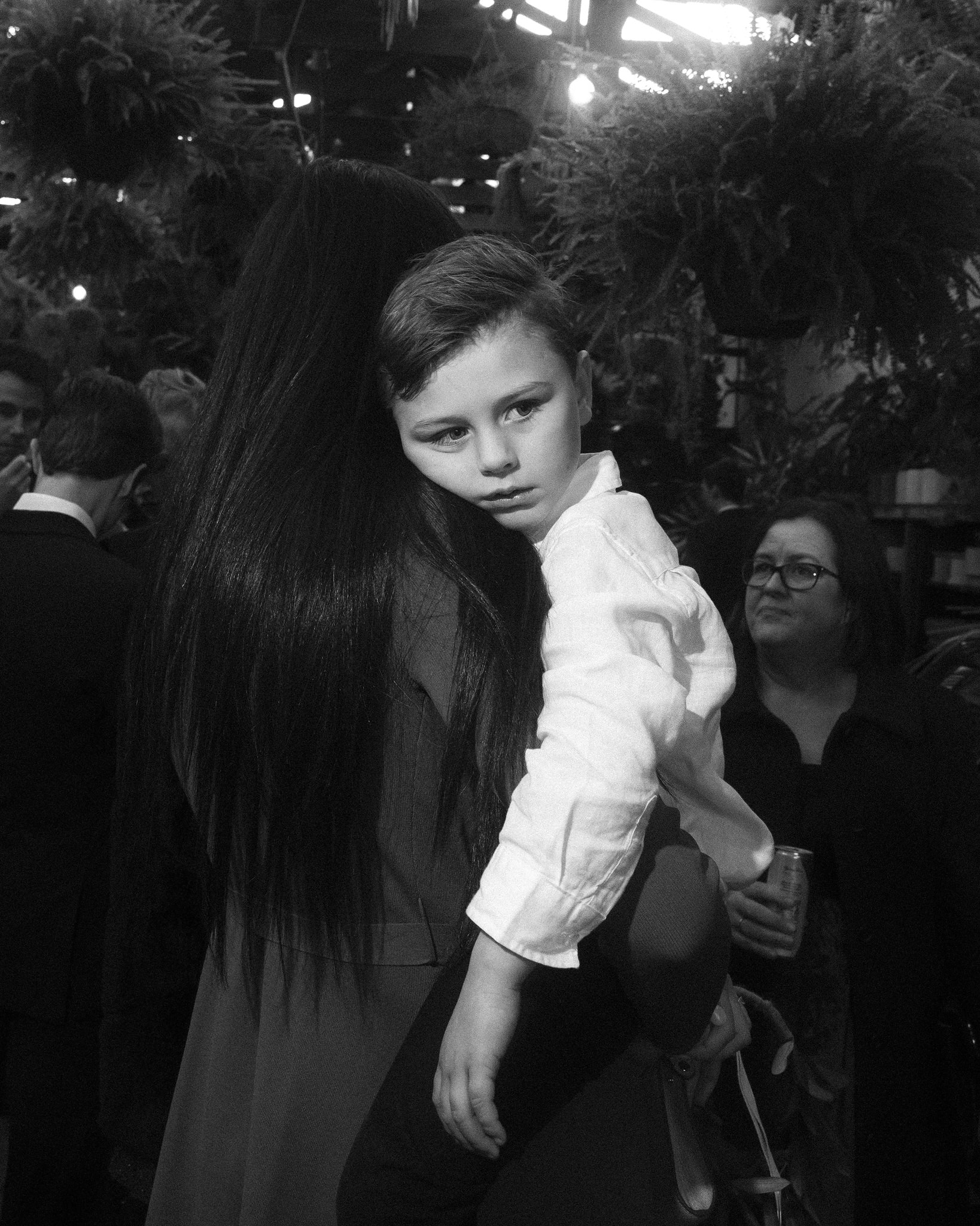 The image is a poignant black and white photograph capturing a tender moment at a wedding. A young boy, dressed in a formal white shirt, rests his head on a woman's shoulder, who appears to be his mother. The woman's long, dark hair flows over her back, providing a soft contrast to the boy's solemn expression and thoughtful gaze directed towards the camera. They are at a wedding reception, surrounded by lush greenery and other guests in the background, which contributes to a festive yet intimate atmosphere. The woman's gentle hold on the boy and his serene posture suggest a moment of comfort and reassurance amidst the bustling celebration, highlighting the deep connection between mother and child during significant family occasions.