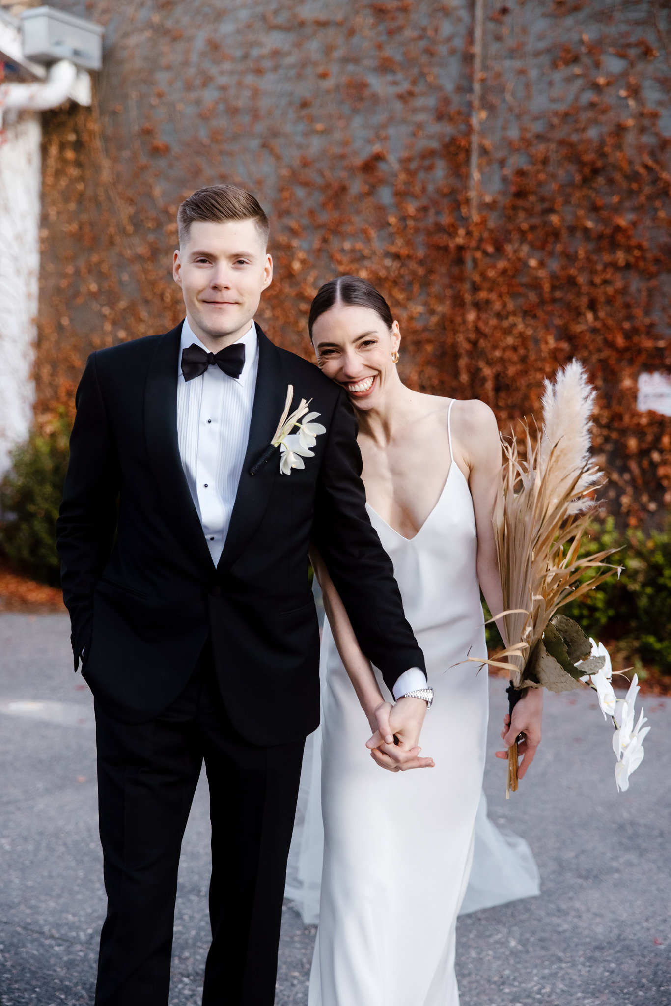 The image captures a joyful and vibrant moment of a bride and groom walking together, set against a backdrop of rusty autumn leaves. The bride, radiant and smiling widely, clutches a unique bouquet featuring white orchids and dried pampas grass. She is dressed in a sleek, elegant white gown with a deep V-neckline. The groom, looking dapper in a classic black tuxedo with a bow tie and a boutonniere that matches the bride's bouquet, shares her joy, holding her hand as they walk. Their expressions and the casual, intimate way they hold hands convey a sense of happiness and shared excitement. The autumn leaves in the background add a rich, colorful texture to the scene, enhancing the festive and romantic atmosphere of the photograph.