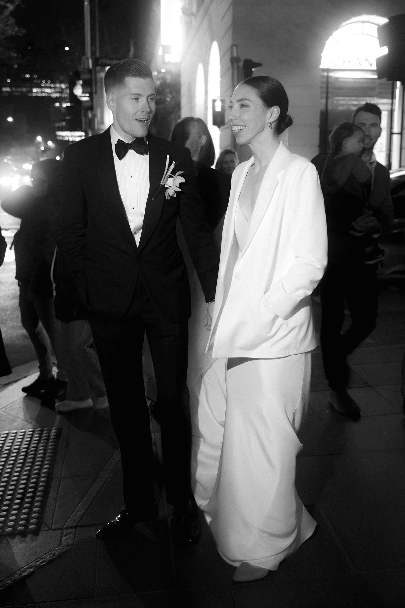The image is a black and white photograph capturing a bride and groom walking together at night, illuminated by the soft glow of city lights. The bride is stylishly dressed in a modern white suit with a tailored jacket and wide-legged trousers, complementing her sleek, pulled-back hair and radiant smile. The groom, in a classic black tuxedo with a bow tie, looks at the bride with an expression of admiration and joy. The couple's relaxed and happy demeanor as they stroll suggests a moment of shared happiness amidst the bustling urban environment. The background features blurred city lights and passersby, focusing the viewer's attention on the couple's intimate interaction. This photograph beautifully blends contemporary wedding fashion with the dynamic energy of a nighttime urban setting.
