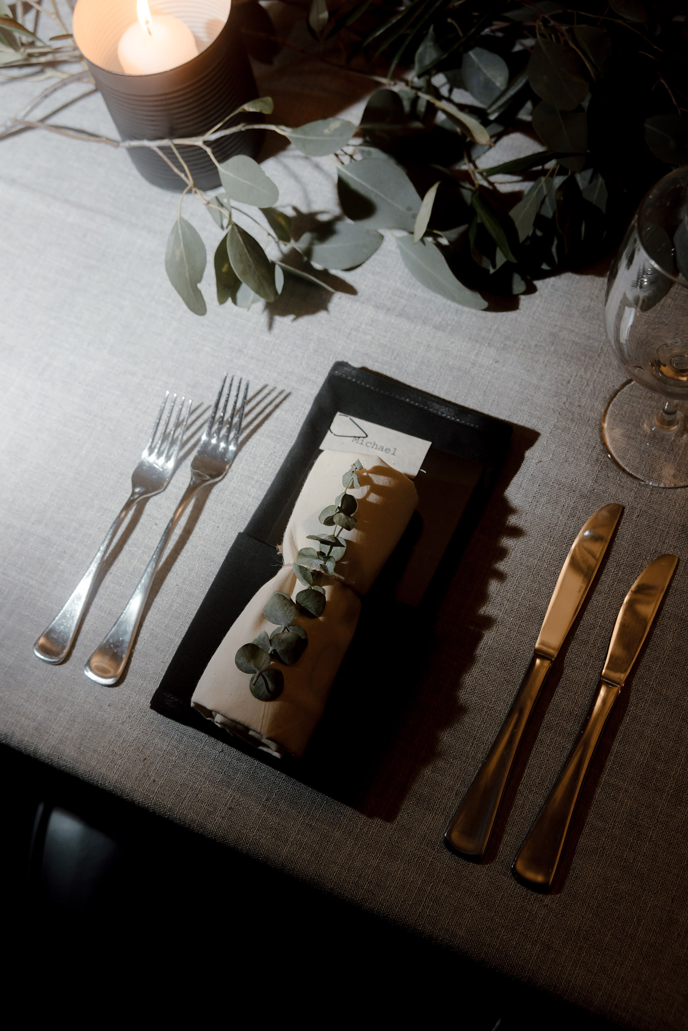 The image features an elegantly set wedding table detail, capturing a sophisticated and serene dining atmosphere. A napkin, delicately wrapped with a rustic string and adorned with eucalyptus leaves, rests on a sleek black charger plate, creating a beautiful contrast with the light, textured linen tablecloth. Beside the plate are polished silver and gold cutlery, adding a touch of luxury. A lit candle in a black holder casts a warm glow, enhancing the cozy, intimate vibe of the setting. The soft, diffused lighting highlights the simplicity and elegance of the table decor, perfectly suited for a modern wedding celebration. This setting not only provides a visual treat but also complements the overall sophisticated and nature-inspired theme of the event.
