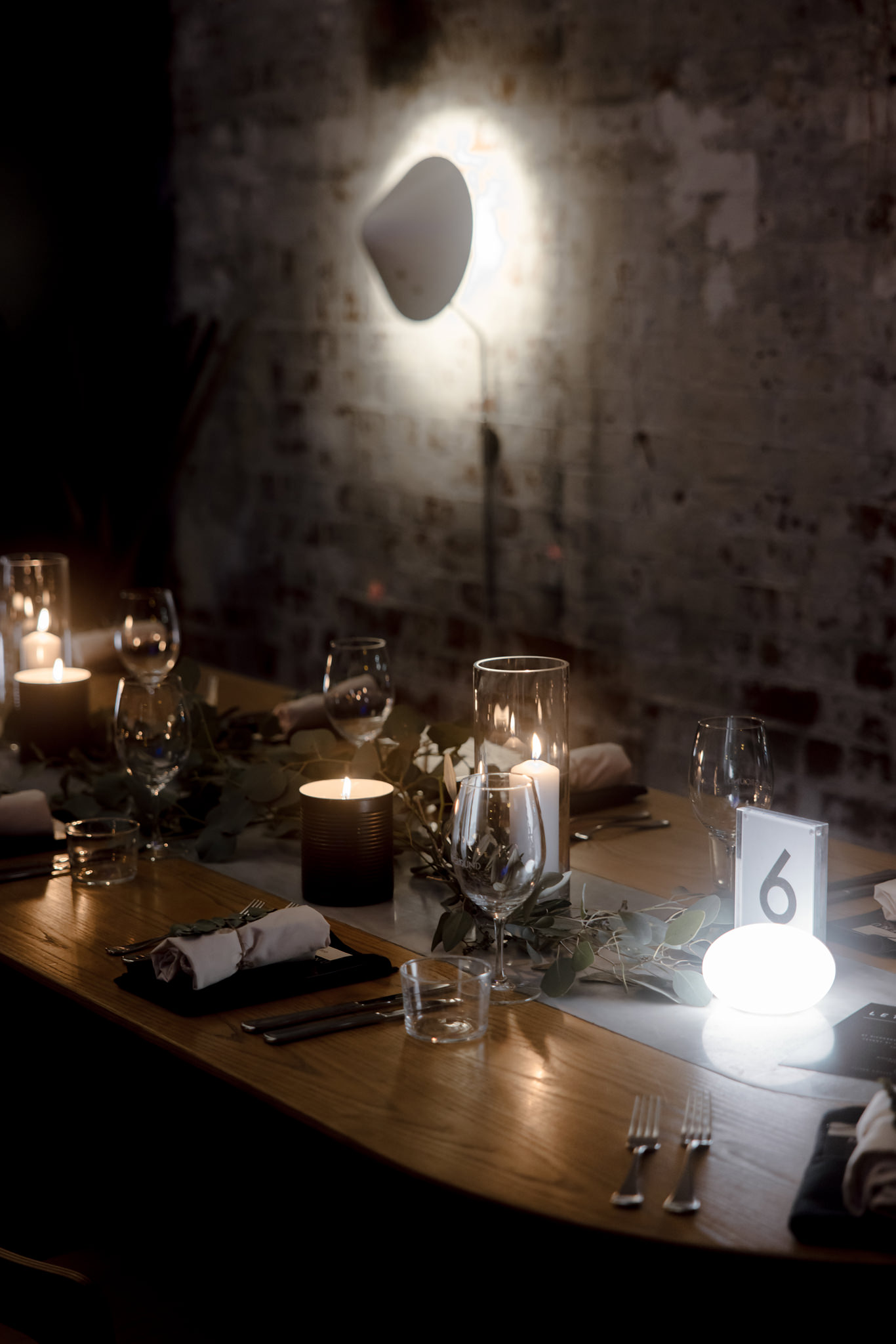 The image is a beautifully composed photograph of a wedding reception table set in a moody and atmospheric venue. The scene is lit by a mix of soft candlelight and a unique wall-mounted light casting a distinct shape on the rustic brick background, which adds texture and an industrial chic vibe to the setting. The wooden table is elegantly adorned with simple yet stylish elements: glassware, candles in dark holders, and fresh greenery strewn across the center, creating a natural, understated look. A table number "6" glows softly in a small illuminated stand, providing a modern touch amidst the traditional setting. This table setup captures the essence of a contemporary wedding that blends romantic and urban aesthetics, offering a warm and inviting atmosphere for the guests.