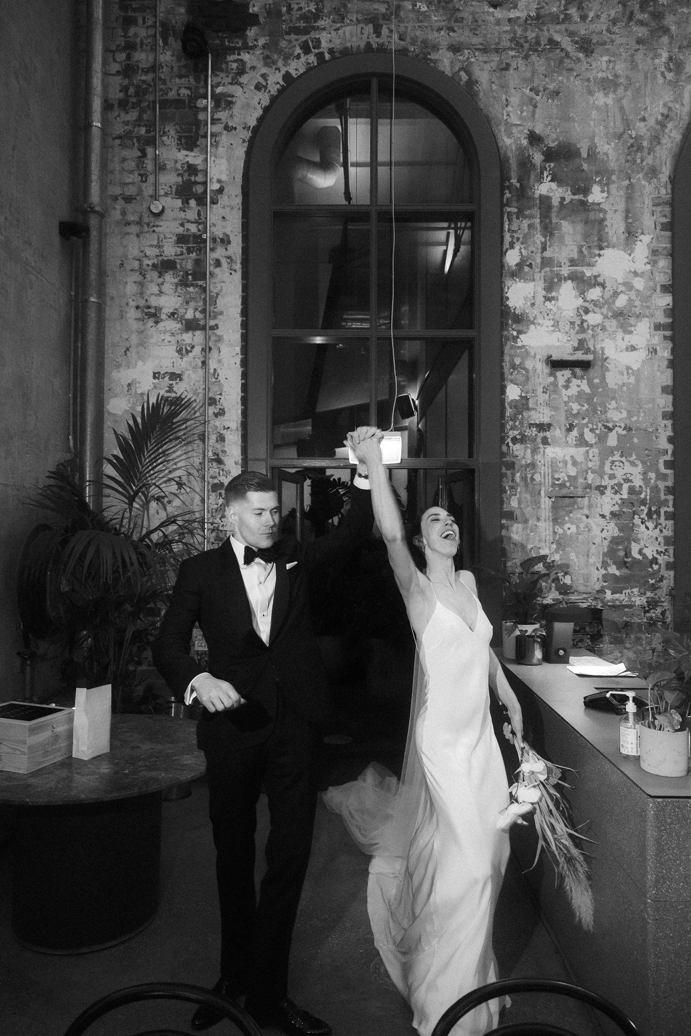 Samantha and Michael stand together in a sunlit corridor of the Higher Ground wedding venue. She wears a flowing white gown, and he is in a sleek, dark suit. They are framed by the rustic, industrial walls, which blend modern wedding elegance with the building's historic charm.