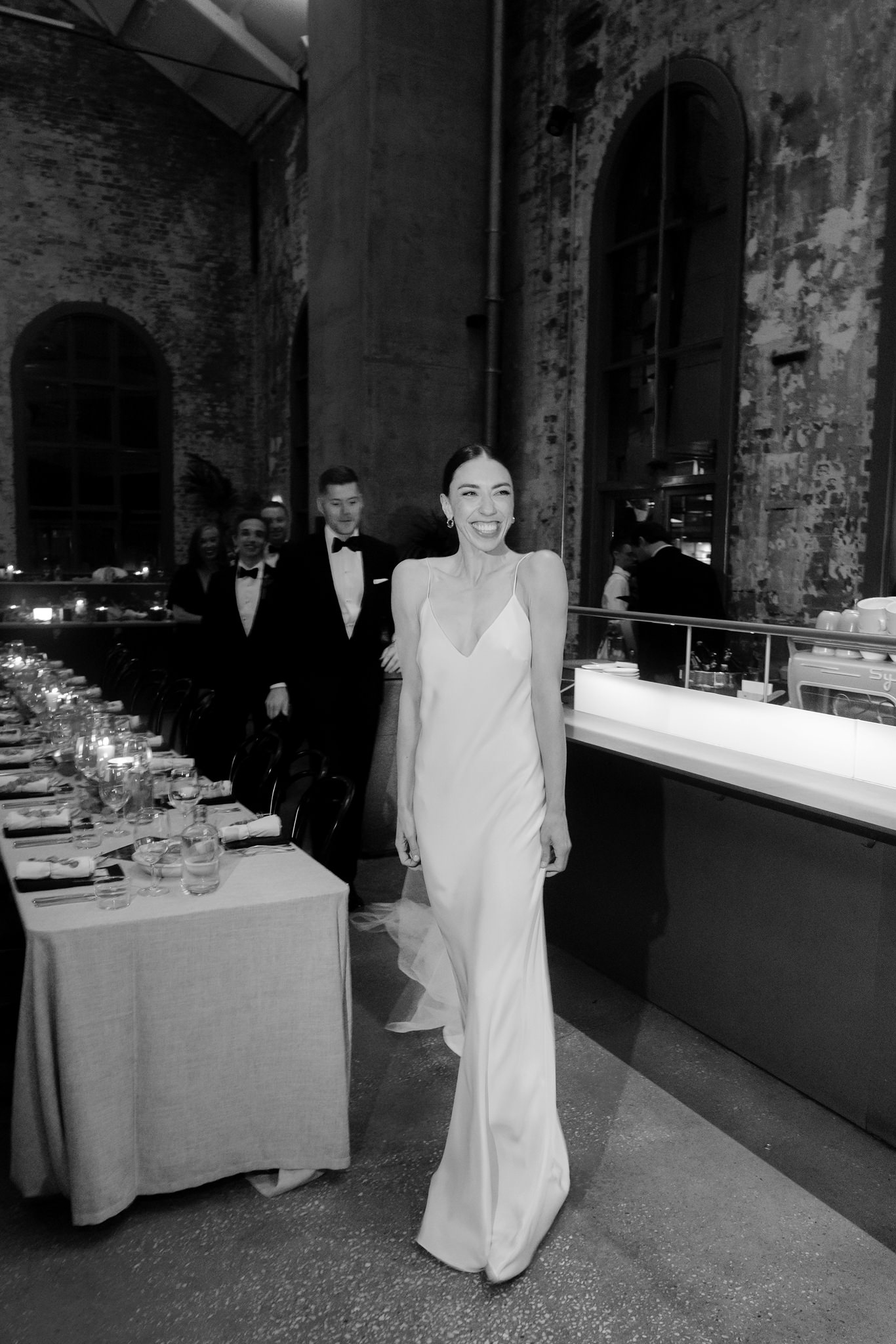 In this captivating black and white wedding photograph, the bride is the center of attention as she strides joyfully through the venue. She wears a sleek, form-fitting white gown with thin straps and a plunging neckline, complemented by a delicate, flowing veil trailing behind her. Her face lights up with a radiant smile, embodying the happiness of her special day. Behind her, the groom and a group of well-dressed guests follow, all sharing in the festive spirit. The rustic elegance of the venue, characterized by its tall arched windows and exposed brick walls, provides a stunning backdrop, enhancing the timeless and elegant atmosphere of the moment. The image beautifully captures the exuberance and elegance of the wedding celebration, set in a venue that combines historic charm with contemporary style.
