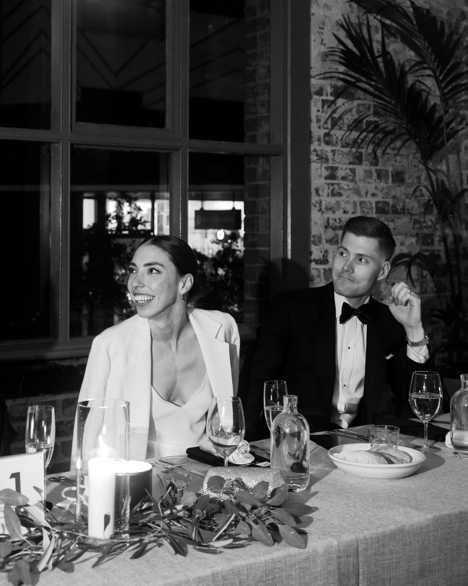 In this elegant black and white wedding photo, the bride and groom are captured seated at their reception table, reflecting a moment of joyful interaction. The bride, to the left, is dressed in a chic white blazer over her wedding gown, laughing openly and looking away from the camera, her expression one of genuine happiness. The groom, to the right, glances towards her with a playful smirk, wearing a classic black tuxedo and bow tie. They are surrounded by an intimate table setting featuring candles, wine glasses, and a decorative arrangement of leaves and greenery that enhances the rustic charm of the brick wall and large pane windows behind them. This image beautifully encapsulates a candid, heartfelt moment of the couple's wedding reception, set against a backdrop that blends urban and natural elements.