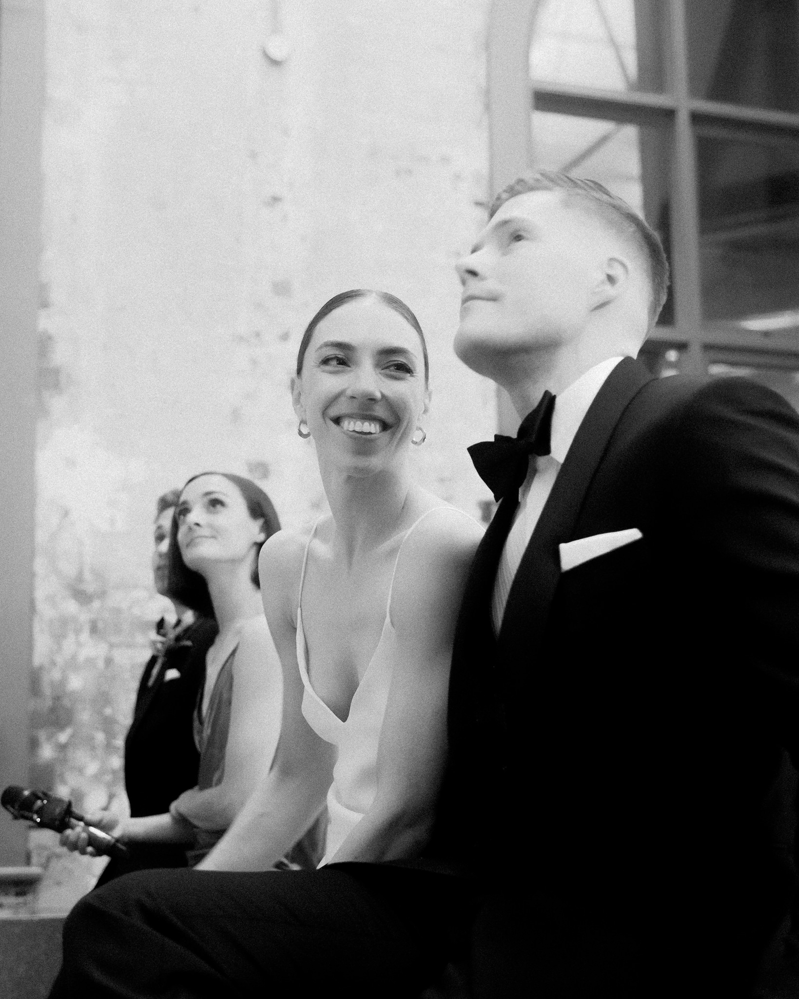 This photograph captures a beautiful moment at a wedding, presented in black and white for a timeless feel. It shows the bride and groom, along with another female guest, all looking upwards, seemingly captivated by a speech or event out of the frame. The bride, centrally located, sports a broad, joyful smile, her face radiating happiness. She is dressed in a sleek, low-cut wedding gown that enhances her elegance, while the groom, to her right, appears in a sharp tuxedo and bow tie, his expression one of attentive admiration. The other female guest, to the left of the bride, also gazes upward, her expression thoughtful and engaged. The backdrop features the rustic charm of a distressed brick wall and large windows, adding to the ambiance of the venue. This image beautifully conveys the joy and collective engagement of the guests at this significant moment, highlighting the emotional resonance of the occasion.