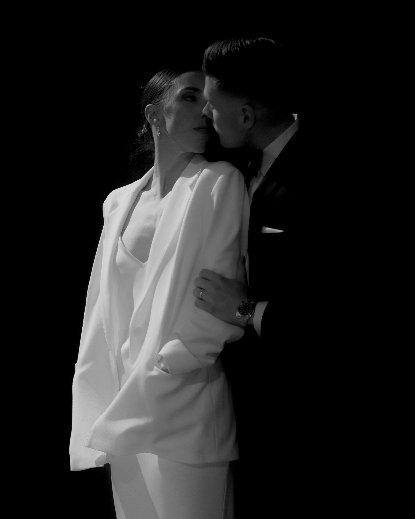 This evocative black and white image captures a deeply intimate moment between a bride and groom during their wedding. The bride, donned in a sleek white suit jacket over her dress, looks tenderly at the groom who leans in close to her, their faces nearly touching, suggesting an imminent kiss. The groom, in a sharp black tuxedo, embraces the bride gently around her waist. Their expressions are serene and filled with affection, highlighted against the stark black background that enhances the emotional and dramatic quality of the scene. This photograph beautifully conveys the love and connection between the couple, encapsulated in a quiet, private moment amidst the celebration.
