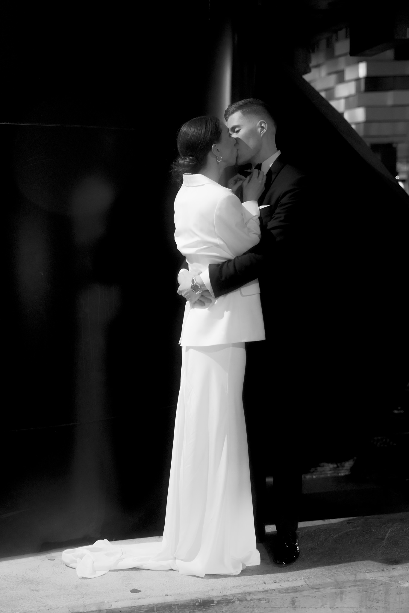 This black and white wedding photograph captures a tender moment between the bride and groom as they share a kiss on a city street at night. The bride, clad in a stylish white suit with a long, flowing train, embraces the groom, who is dressed in a classic black tuxedo. The scene is lit by the soft glow of streetlights, creating a dramatic contrast between the dark background and the illuminated figures of the couple. The setting includes blurred architectural details and a slightly visible urban environment, adding a sense of romance and intimacy to this special moment. The bride's joy is palpable as she smiles during the kiss, and the groom gently holds her face, making this image a timeless portrayal of love and commitment.