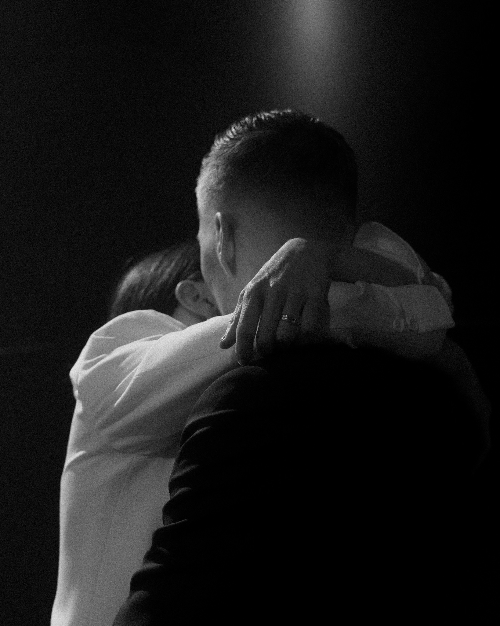 This intimate black and white wedding photograph captures a close-up moment between the bride and groom as they embrace. The focus is on the groom's back and head, showing his short, neatly styled hair and the top of his black suit. The bride, visible from behind, wraps her arms around the groom's neck, her hand gently resting on his shoulder. Her engagement and wedding rings are visible, adding a touch of sparkle to the image. The lighting highlights their upper bodies, creating a soft glow around them while the background fades into darkness, emphasizing the closeness and emotion of their embrace. This powerful image conveys a sense of tenderness and the deep connection shared by the couple.
