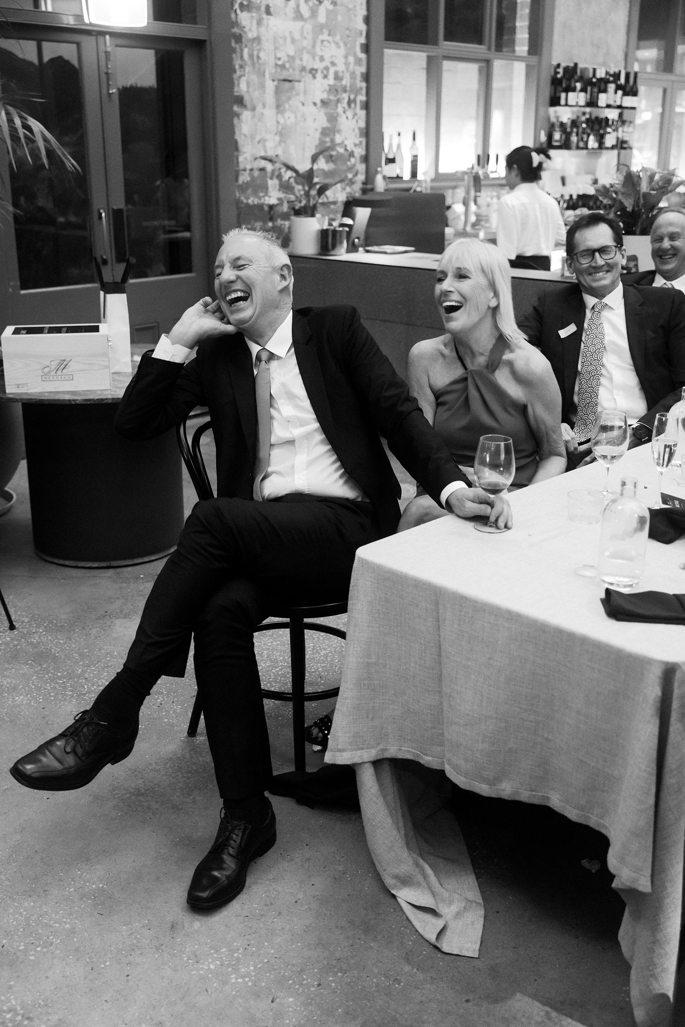 This black and white photograph captures a moment of genuine joy at a wedding reception held at Higher Ground. The image shows a man and a woman seated at a dining table, both dressed in formal attire. The man, wearing a classic black suit, is leaning back in his chair laughing heartily, his hand to his ear, while the woman next to him, in an elegant off-shoulder dress, laughs openly with her hand on his shoulder. Their expressions of unguarded laughter, combined with the festive environment, characterized by a casually elegant setting with rustic brick walls and modern decor in the background, evoke a sense of warmth and celebration.