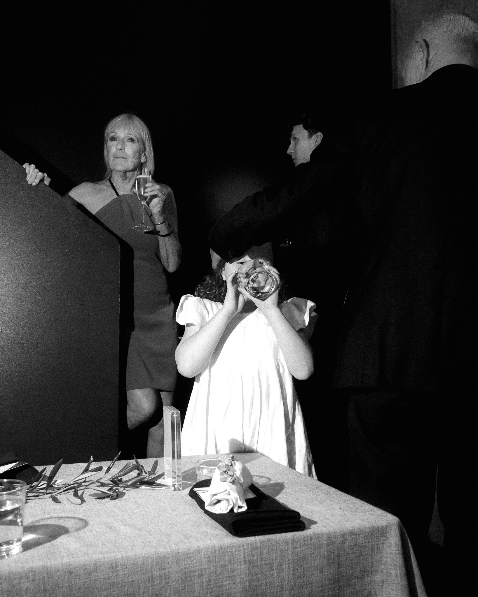 This black and white photograph from a wedding reception at Higher Ground features a candid scene among guests. In the foreground, a young girl in a white dress drinks playfully from a clear bowl, completely oblivious to her surroundings. Behind her, a mature woman, elegantly dressed in a one-shoulder gown, stands holding a glass of wine, observing the festivities with a subtle smile. Beside her, a young man in a dark suit leans over a partition, watching the events unfold. The setting has a minimalist and modern aesthetic, highlighted by the stark contrast of the black background and the illuminated figures, adding a dramatic and intimate ambiance to the scene.