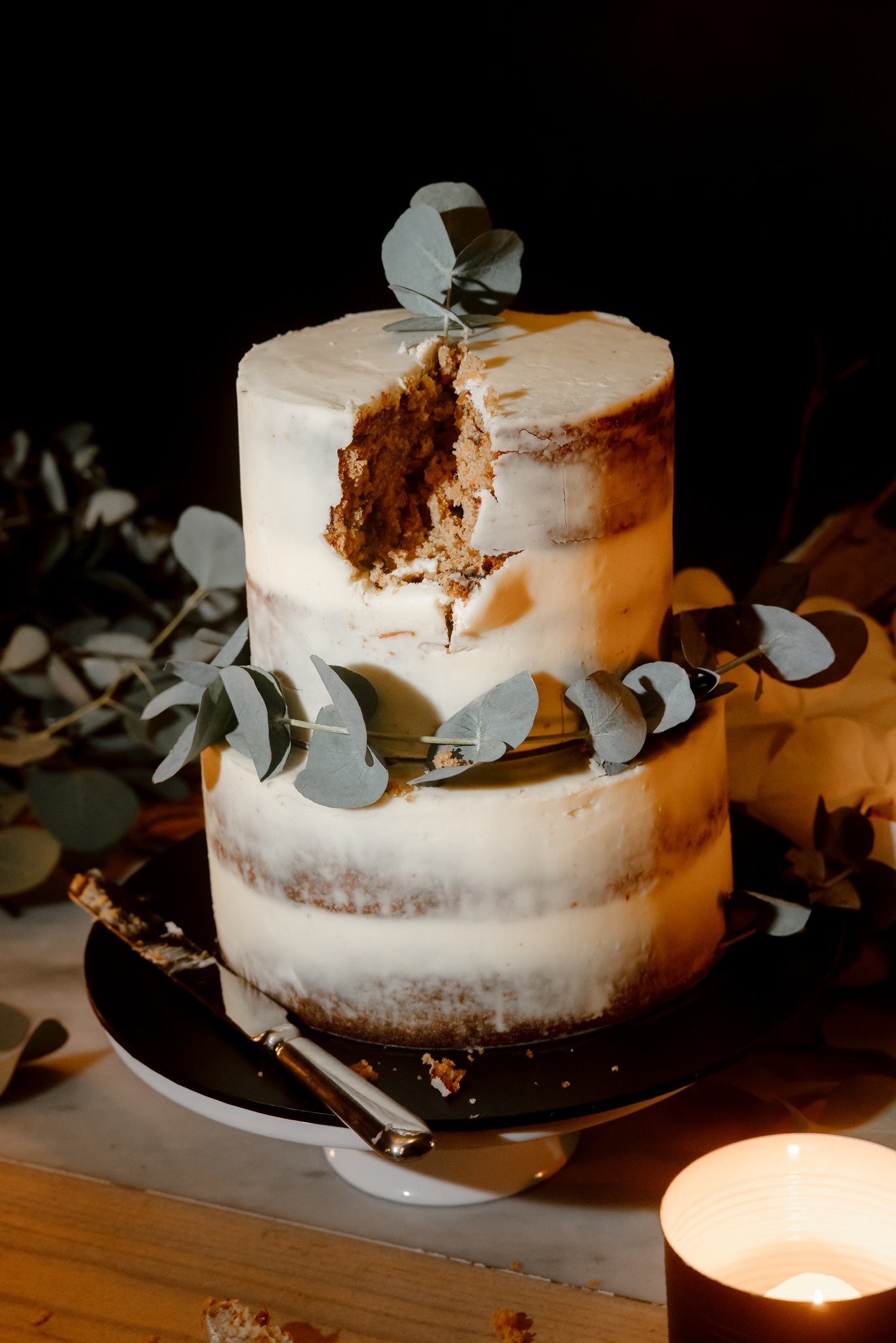 This elegant photograph from a wedding reception features a two-tiered wedding cake. The cake is partially sliced, revealing its rich, moist carrot cake interior, contrasted against the smooth, ivory-colored icing. Decorated simply with a few sprigs of silver dollar eucalyptus, the cake is set on a sleek black plate. The warm, intimate lighting enhances the rustic charm of the cake, with a softly glowing candle in the foreground adding a touch of romantic ambiance. The dark background and minimal decoration focus the attention on the creamy texture and the artisanal look of the cake, embodying a modern yet timeless wedding aesthetic.