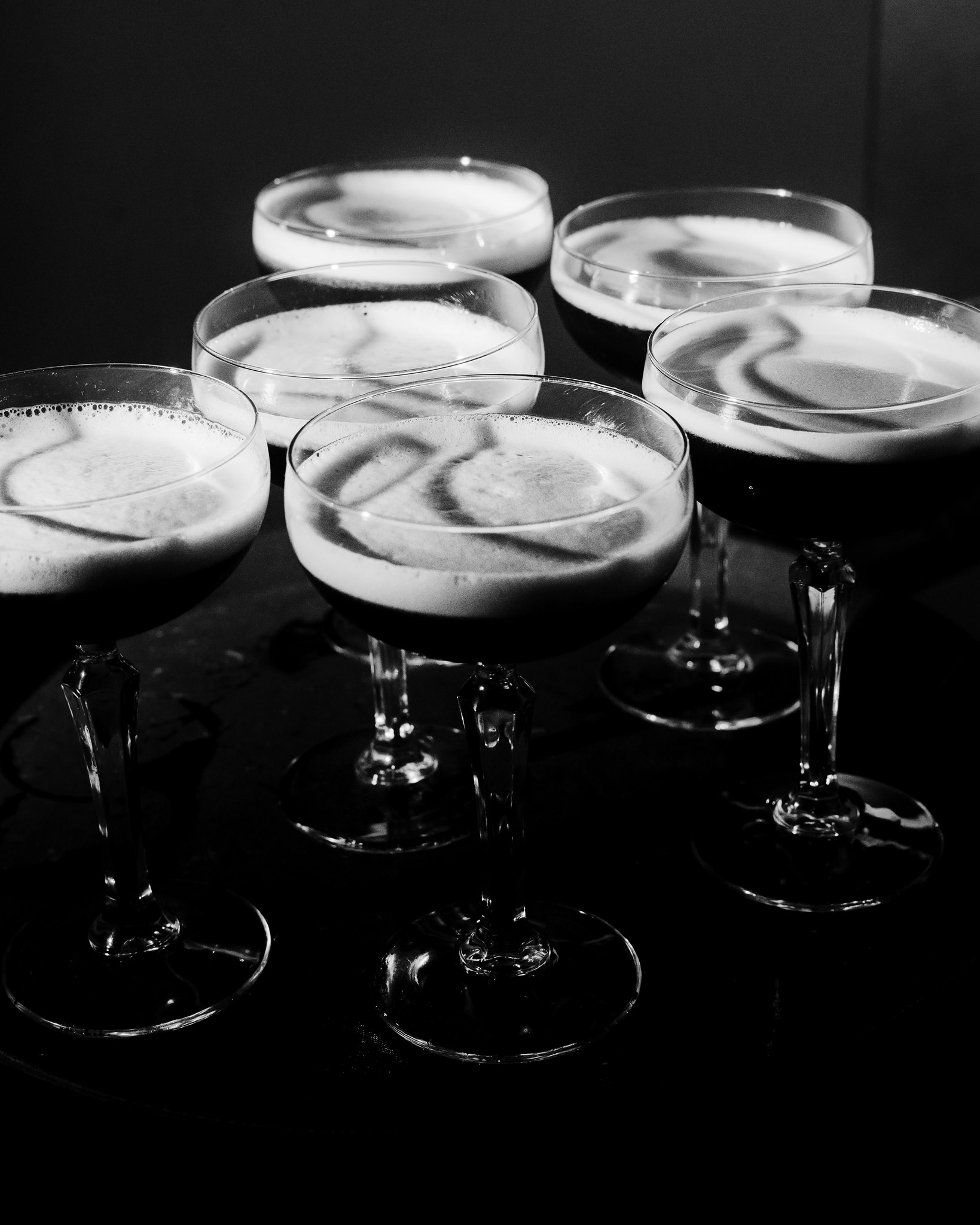 This monochrome photograph captures a sophisticated display of five cocktail glasses, each filled with a dark, frothy beverage, likely a signature cocktail from a wedding event. The glasses are of the coupe style, known for their broad, shallow bowls and elegant stems, arranged closely together on a glossy surface that reflects their silhouettes. The lighting is dramatic, focusing on the creamy froth and delicate patterns created by the foam on the surface of the drinks, enhancing the textures and contours of the glasses. This image conveys a sense of contemporary elegance and the joyous celebratory atmosphere of a wedding, emphasizing a moment of indulgence and festivity.