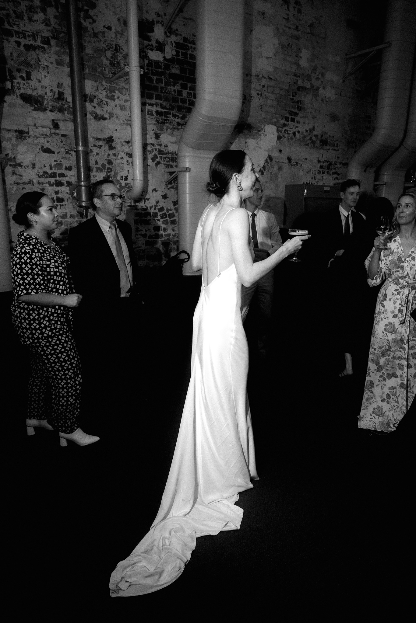 This black-and-white photograph captures a candid moment at a wedding, featuring a bride engaging with guests during the reception. The bride, dressed in a sleek, floor-length gown with a deep V-back and a long flowing train, stands holding a microphone, mid-speech or toast. Her posture and facial expression convey joy and animation as she interacts with her audience. Surrounding her are several guests in semi-formal attire, attentively listening and holding drinks. The backdrop features the raw, textured surface of an old brick wall with exposed pipes, contributing to the industrial chic ambience of the venue. This image vividly portrays a lively, elegant wedding celebration.
