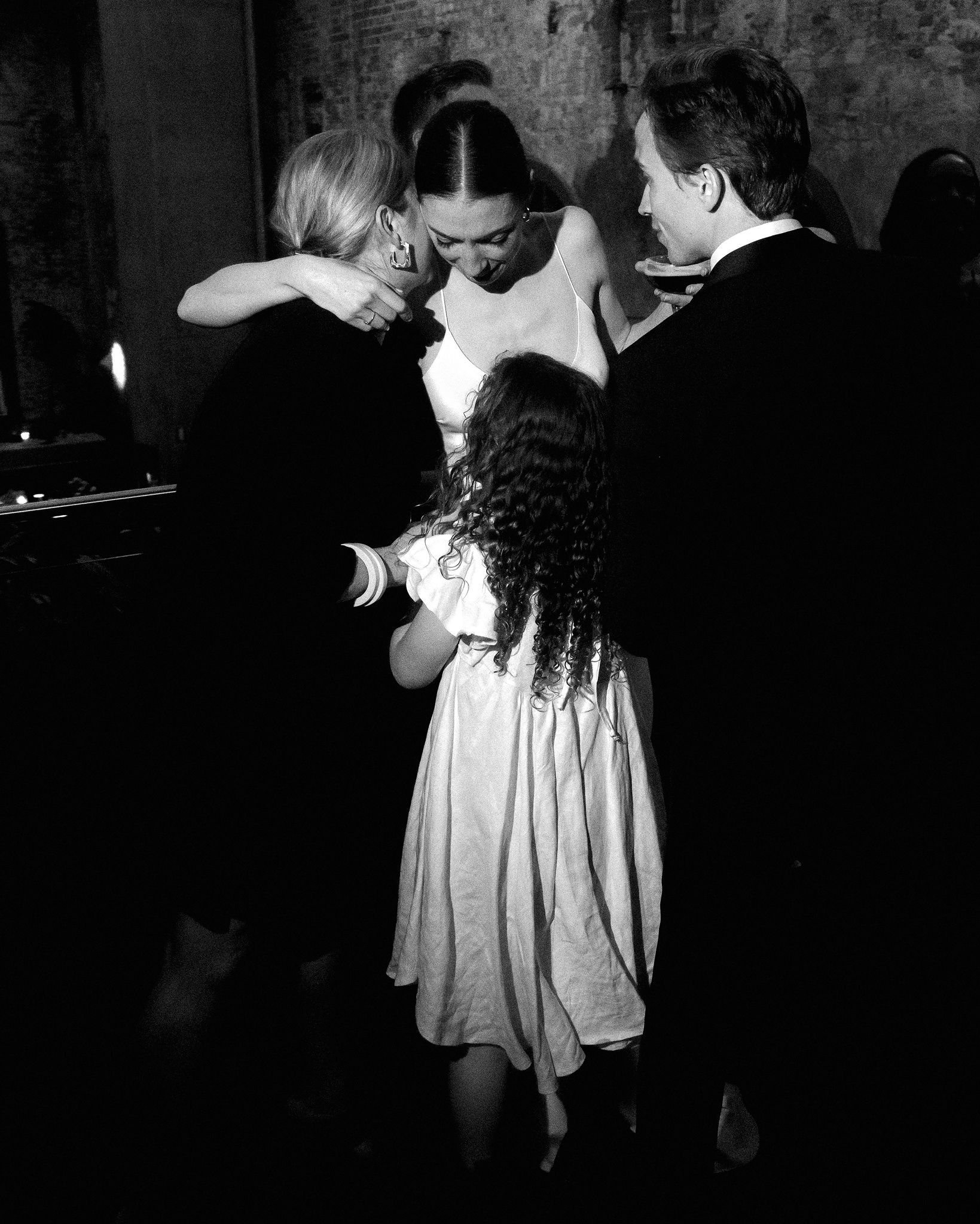 This evocative black-and-white photograph captures a tender moment during a wedding reception, showing the bride in a heartfelt embrace with a group of guests. The bride, dressed in an elegant white gown with a simple neckline, leans forward to hug a woman with blonde hair, who is partially obscured by the embrace. A young girl with curly hair, dressed in a white dress, looks on intently, standing in front of a man in a suit, who is also engaging in the moment. The rustic charm of the venue is evident in the background, highlighted by the rough, exposed brick walls, adding a warm and intimate atmosphere to the scene. This image beautifully encapsulates the joy and closeness of the wedding celebration.