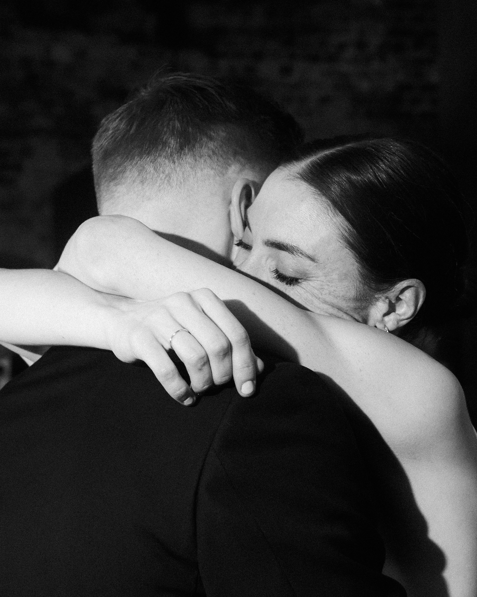 This intimate black-and-white photo captures a close embrace between a bride and groom at their wedding. The groom's back is toward the camera, his head slightly turned, while the bride leans into him, her face pressed against his shoulder, eyes closed, exuding a sense of peace and contentment. The bride's hand is gently placed on the groom's arm, showcasing her engagement ring. Their embrace is framed against a softly blurred background, featuring the rustic texture of an old brick wall, which highlights the tender moment shared between the couple. This image beautifully conveys the emotion and closeness of their special day.