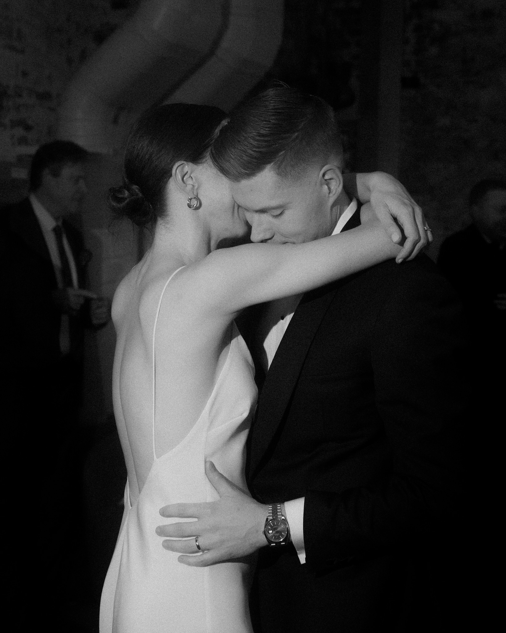 This black-and-white wedding photograph captures a tender moment between the bride and groom. Positioned in a soft embrace, the groom gently rests his forehead against the bride's as she lovingly wraps her arms around his neck. The bride's back is exposed by the elegant design of her dress, adding a touch of delicate sophistication to the scene. Their eyes are closed, highlighting a moment of tranquility and intimate connection amidst the bustling backdrop of guests and soft lighting that gently illuminates the contours of their faces and attire. This image radiates the emotion and closeness of their wedding day, set against the rustic charm of a brick-walled venue.