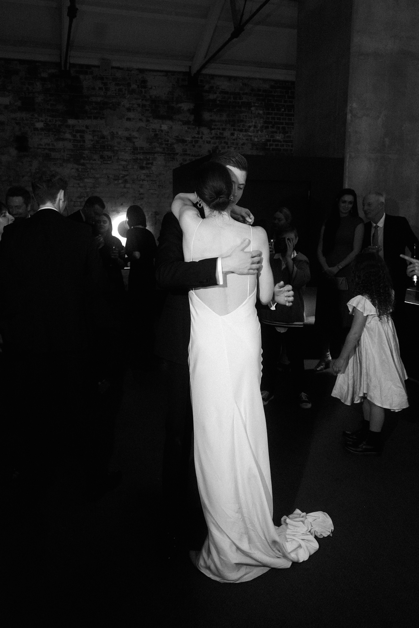 This black-and-white wedding photograph beautifully captures an intimate embrace between the bride and groom during their reception. The bride, dressed in a sleek, open-back gown, wraps her arms around the groom, who reciprocates with a warm embrace. The couple is surrounded by guests who are engaging in conversations and enjoying the festive atmosphere, with a young girl in a white dress visible in the foreground. The soft, ambient lighting enhances the romantic and tender mood of the scene, while the rustic brick walls of the venue add a textured, historic feel to the background. This image eloquently portrays a moment of love and celebration amidst a joyful gathering.