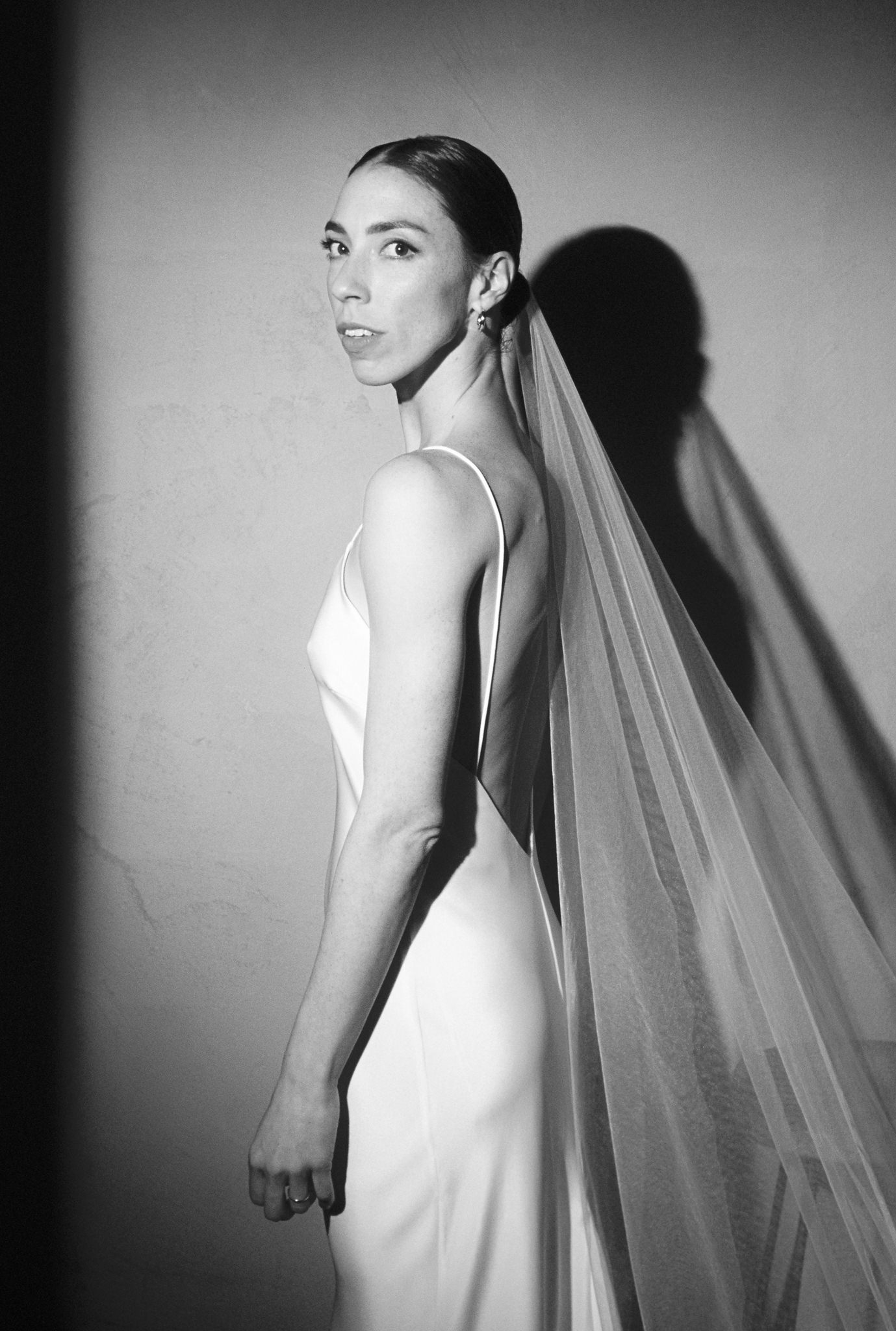 This striking black-and-white wedding portrait features the bride in an elegant, minimalist gown with a slim silhouette and spaghetti straps. She stands against a simple, unadorned wall, turning her head slightly to look over her shoulder at the camera, her expression serene and confident. Her sleek hair is pulled back tightly, accentuating her strong facial features and the bold, geometric earrings that complement her modern look. A delicate veil extends from her head, casting a soft shadow that merges with her own on the wall behind her, creating an ethereal and dramatic visual. The simplicity of the background and the stark lighting focus all attention on her, highlighting her graceful posture and the understated elegance of her attire.