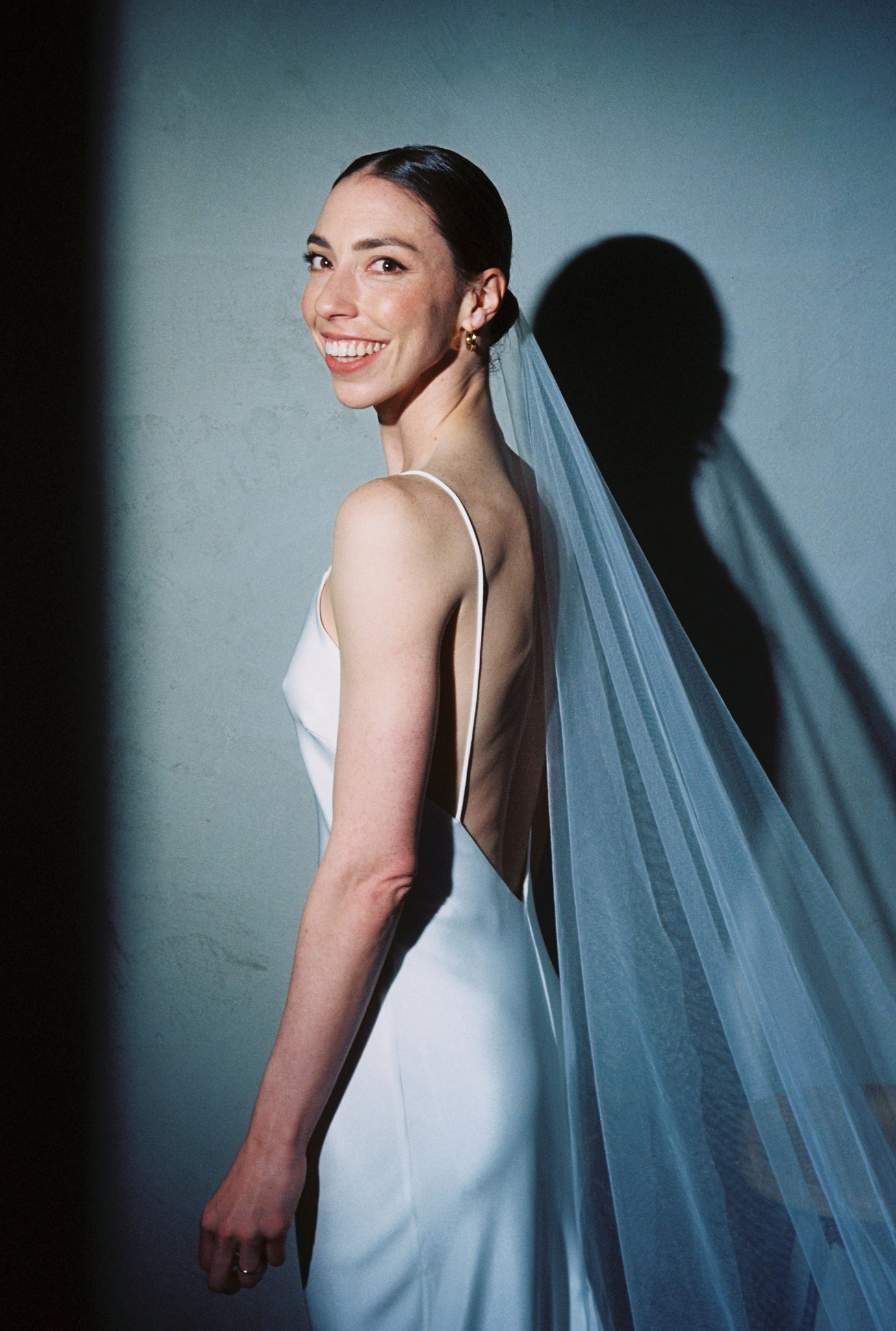 This captivating wedding portrait captures the bride in a joyous moment, her smile bright and welcoming as she looks over her shoulder. She wears an elegant, form-fitting white gown with a deep, plunging back and delicate spaghetti straps that highlight her slender frame. Her long, sheer veil cascades gracefully down her back, adding a classic bridal element to the modern style of her dress. The background is a simple, pale blue wall, providing a soft contrast that enhances the natural beauty of her features and the intricate details of her understated, yet sophisticated, earrings. The lighting casts a dramatic shadow behind her, echoing her pose and adding depth to the composition, while her radiant expression conveys a sense of bliss and excitement.