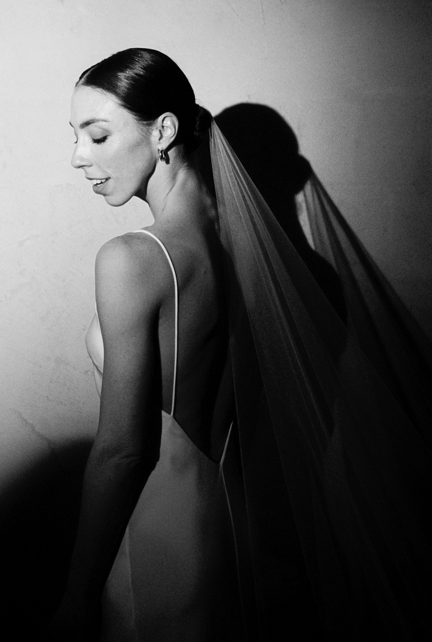 This black and white image captures a serene moment from a wedding, focusing on the bride in profile. She is dressed in a sleek, form-fitting sleeveless gown with a plunging back that highlights the graceful lines of her shoulders and back. Her hair is styled in a smooth, elegant bun, and she wears minimalistic earrings, adding a touch of sophistication. The soft tulle of her long veil trails behind her, casting a dramatic shadow on the simple, unadorned wall beside her. This portrait, set against a minimalist backdrop, emphasizes the bride’s calm demeanor and the elegant simplicity of her wedding attire, capturing a moment of quiet reflection amidst the celebrations.