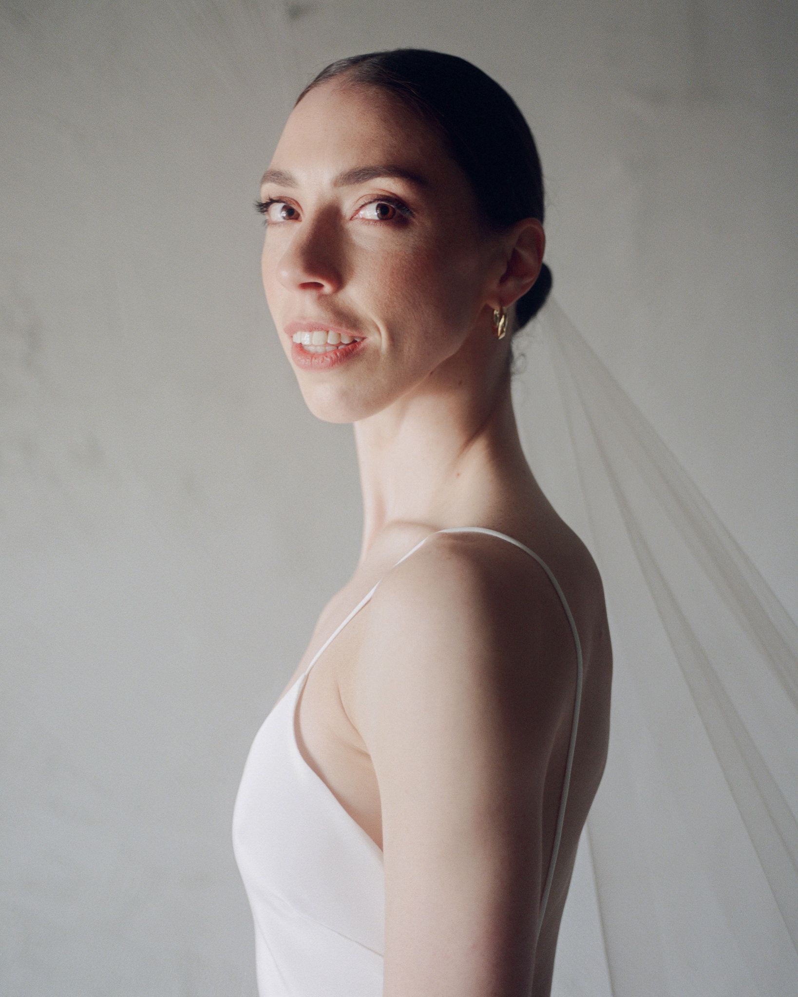This image captures the bride in a serene moment, illuminated by soft natural light. She is portrayed in a three-quarter profile, gazing gently out of the frame with a subtle smile, highlighting her gracefully defined facial features. The bride's elegant updo and delicate earrings complement the minimalist design of her sleek, white gown which features thin straps and a low, rounded back, enhancing her slender silhouette. A sheer, delicate veil cascades from her hair, adding a traditional touch to her modern look. The background is a simple, lightly textured white wall, ensuring all focus is on the bride’s radiant expression and the sophisticated simplicity of her wedding attire. This portrait exudes a sense of calm and joy, typical of a contemplative moment on a special day.