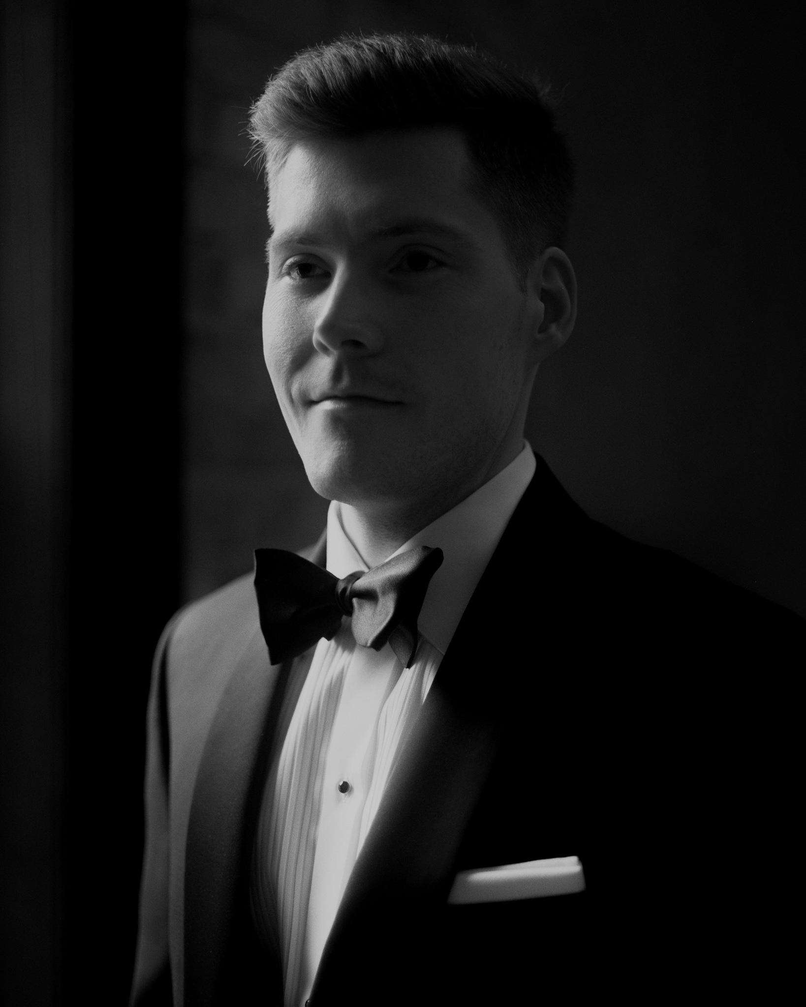 This photograph features a portrait of the groom in a classic black tuxedo with a crisp white shirt and a bow tie, captured in monochrome for a timeless look. His expression is contemplative and calm, gazing slightly off-camera with a serene confidence. The soft, directional lighting gently illuminates one side of his face, enhancing the contours of his features while casting the other side in subtle shadow, creating a dramatic yet intimate atmosphere. The background remains dark, focusing all attention on the groom's dignified and elegant appearance. This image beautifully captures the groom's poised and reflective mood, adding depth to the narrative of the wedding day captured in these images.