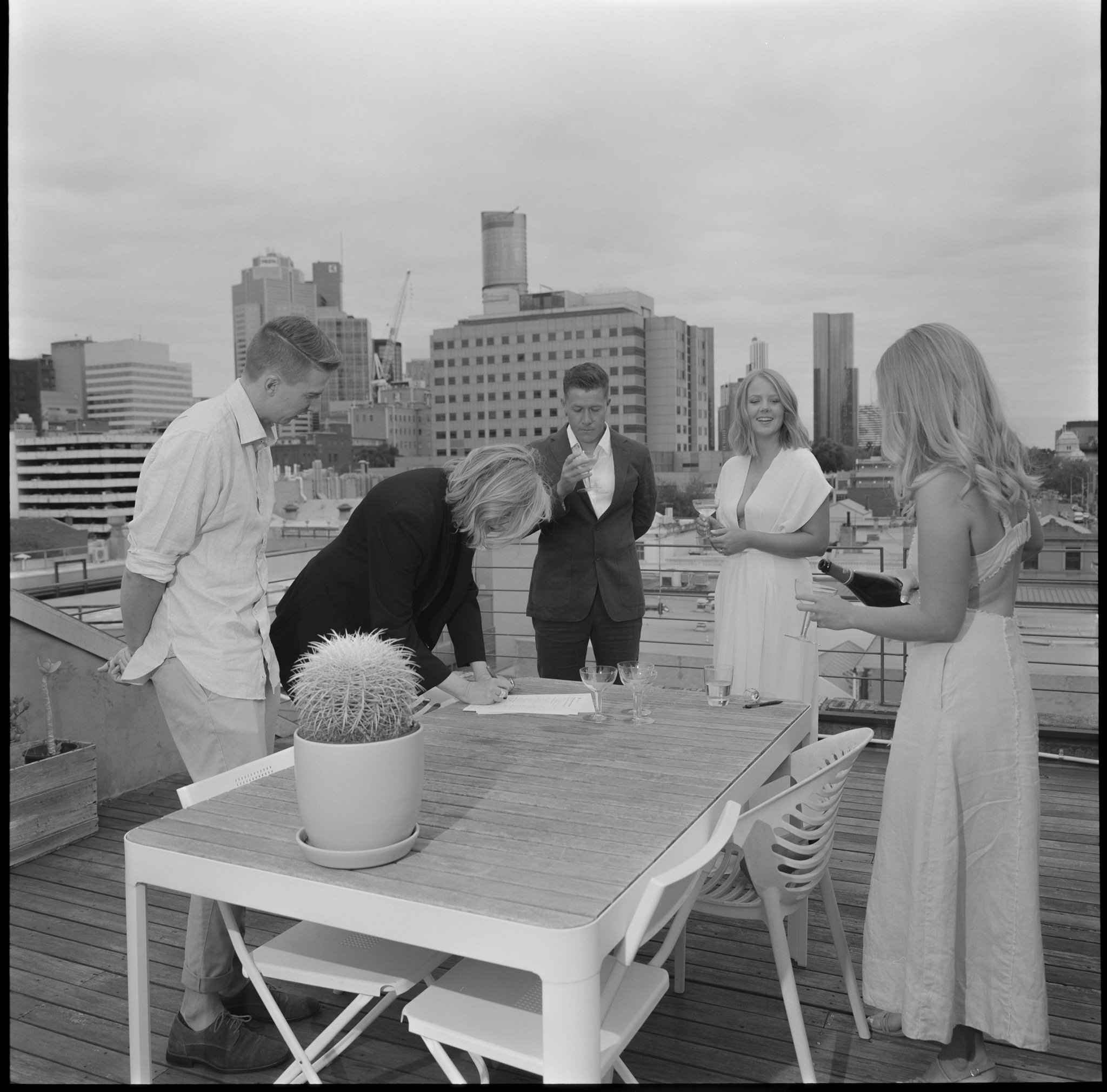 Intimate rooftop wedding ceremony at Recoleta in Melbourne, featuring a couple signing their marriage documents surrounded by close friends and stunning views of the city skyline. The bride, dressed in a minimalist white gown, smiles as the officiant finalizes the paperwork, while friends look on in a relaxed, modern setting. The rooftop's urban atmosphere, complete with chic decor and a view of Melbourne’s iconic skyscrapers, creates a perfect blend of elegance and intimacy. This candid moment captures the essence of a Recoleta wedding in Melbourne—an exclusive, personalized celebration with a backdrop of modern city life.
