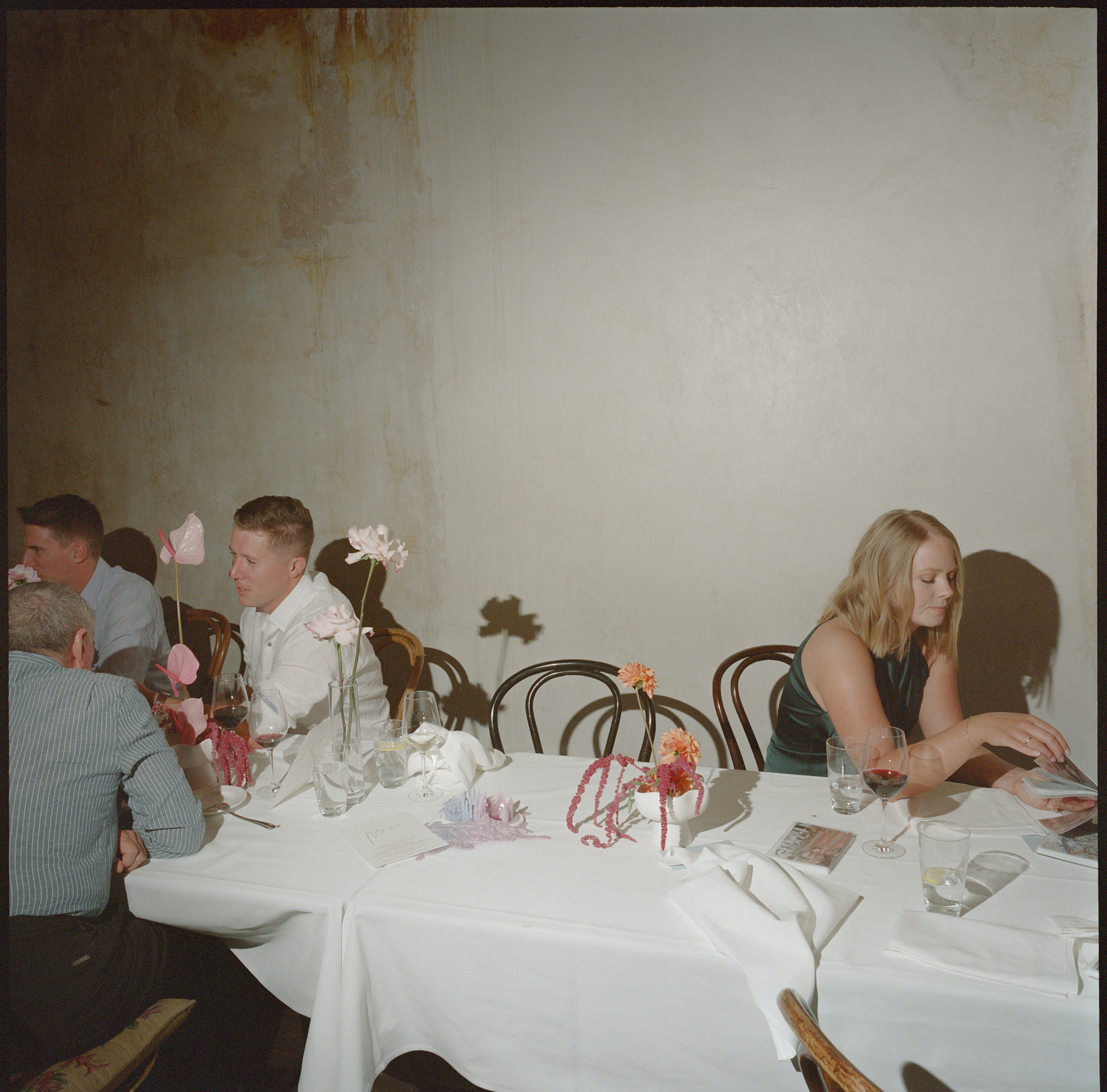 This photograph from a wedding at Recoleta in Melbourne captures a quiet yet thoughtful moment amid the reception. Guests are seated around a long white-clothed table, their conversations intimate and reflective. On the left side of the image, three men sit together, their faces engaged in quiet discussion. One guest, in a white shirt, leans forward with a focused expression, adding to the atmosphere of connection and closeness. The chairs are classic bentwood, lending a timeless touch to the scene.

In contrast, to the right, a woman sits alone, dressed elegantly in a sleeveless dark dress, her attention fully absorbed as she flips through what appears to be a program or card. She is poised and calm, her wine glass half full, suggesting that she's taking a brief respite from the festivities. The lighting is soft and warm, casting gentle shadows on the wall behind her, accentuating the weathered texture of the space, which contrasts with the formal, clean lines of the table.

The table itself is minimally adorned with small yet delicate flower arrangements in soft hues of pinks, oranges, and lavenders, some standing tall in thin vases, their shadows dancing on the wall in the subdued lighting. Scattered across the table are empty glasses, napkins, and bits of decoration, remnants of a celebratory meal, which subtly hint at the lively atmosphere that preceded this quieter moment. The juxtaposition of the guests deep in conversation with the solitary woman flipping through cards brings a serene stillness to the image, encapsulating the multi-faceted nature of weddings—moments of joy, reflection, and connection.