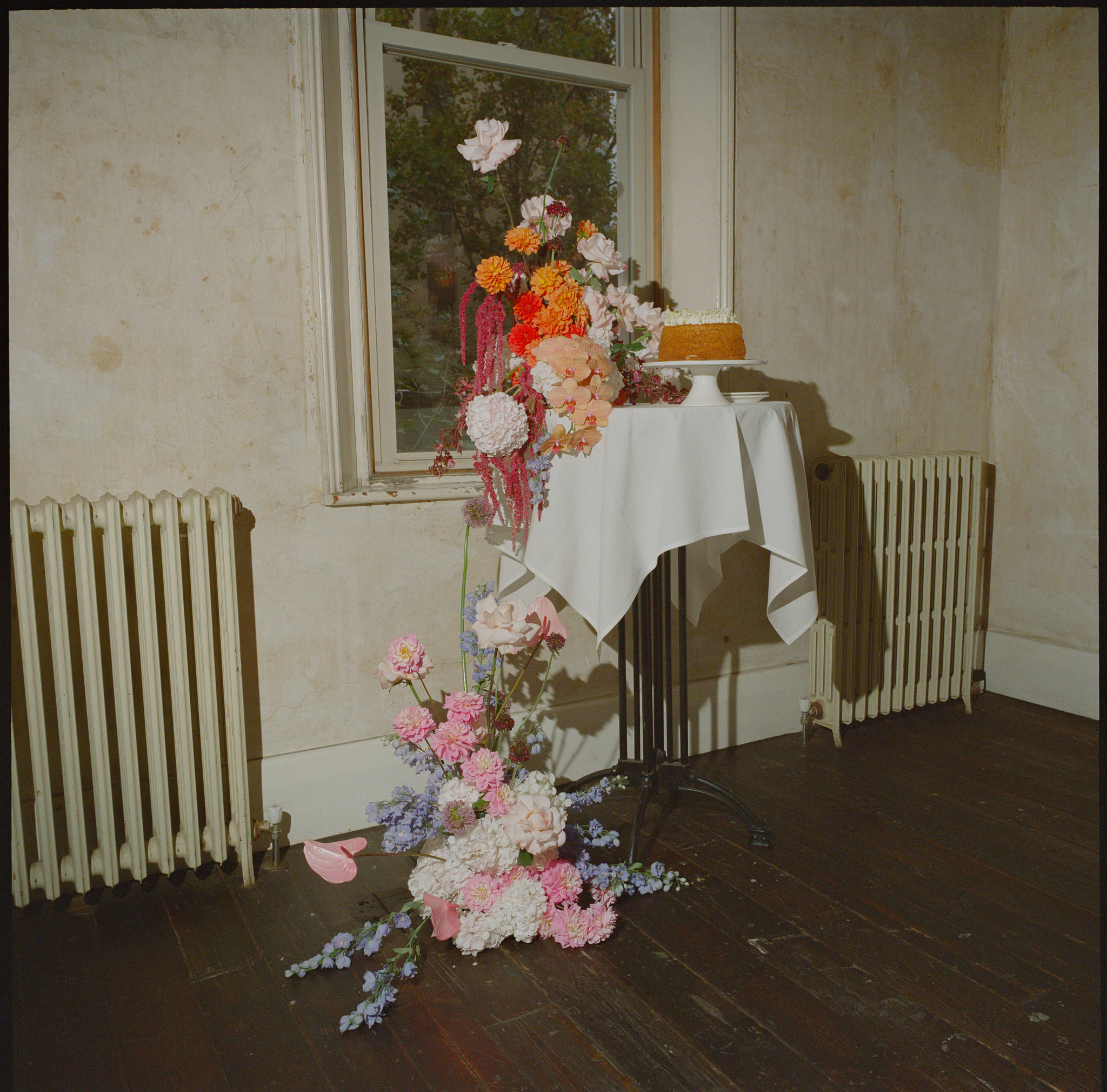 This photograph from a wedding at Recoleta in Melbourne highlights a beautifully arranged cake table set against the backdrop of a rustic, slightly weathered room. The table, covered with a simple white cloth, holds a modest single-tiered cake perched elegantly on a white cake stand. Next to the cake is an opulent floral display that extends from the floor to nearly the height of the window behind it. The arrangement bursts with color, featuring blush pink peonies, vibrant orange zinnias, red hanging amaranthus, soft lavender blooms, and delicate pale pink roses. The flowers cascade down towards the dark wooden floor, adding a vibrant touch of life to the otherwise minimal setting.

The room itself has an understated, vintage charm. Its off-white, aged walls, complete with peeling paint and exposed patches, are complemented by a pair of old-fashioned radiators, further emphasizing the historic, slightly industrial atmosphere. The window offers a glimpse of greenery outside, contrasting with the soft pastel tones and bold florals that dominate the interior scene. The entire setting exudes a mix of old-world charm and modern floral artistry, creating an inviting and romantic atmosphere for the intimate wedding celebration.