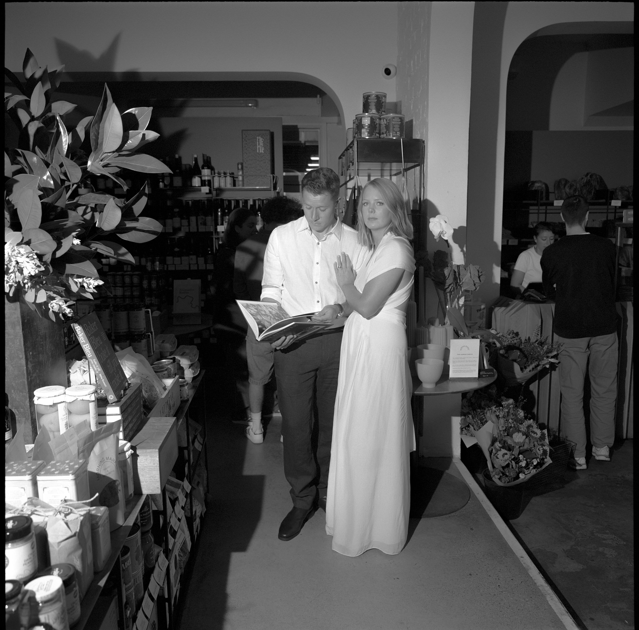 Bride and groom in a candid moment during their Recoleta wedding in Melbourne, captured inside a cozy, boutique market. The couple stands together, with the groom flipping through a book, while the bride rests her hand affectionately on his shoulder. The intimate atmosphere is enhanced by the soft lighting and rustic decor, surrounded by shelves of gourmet products and floral arrangements. This unique setting adds a modern, personalized touch to their Recoleta wedding, blending urban culture with a warm, intimate vibe. The image highlights the couple’s relaxed, elegant style, set against the backdrop of Melbourne's dynamic, eclectic wedding venues.