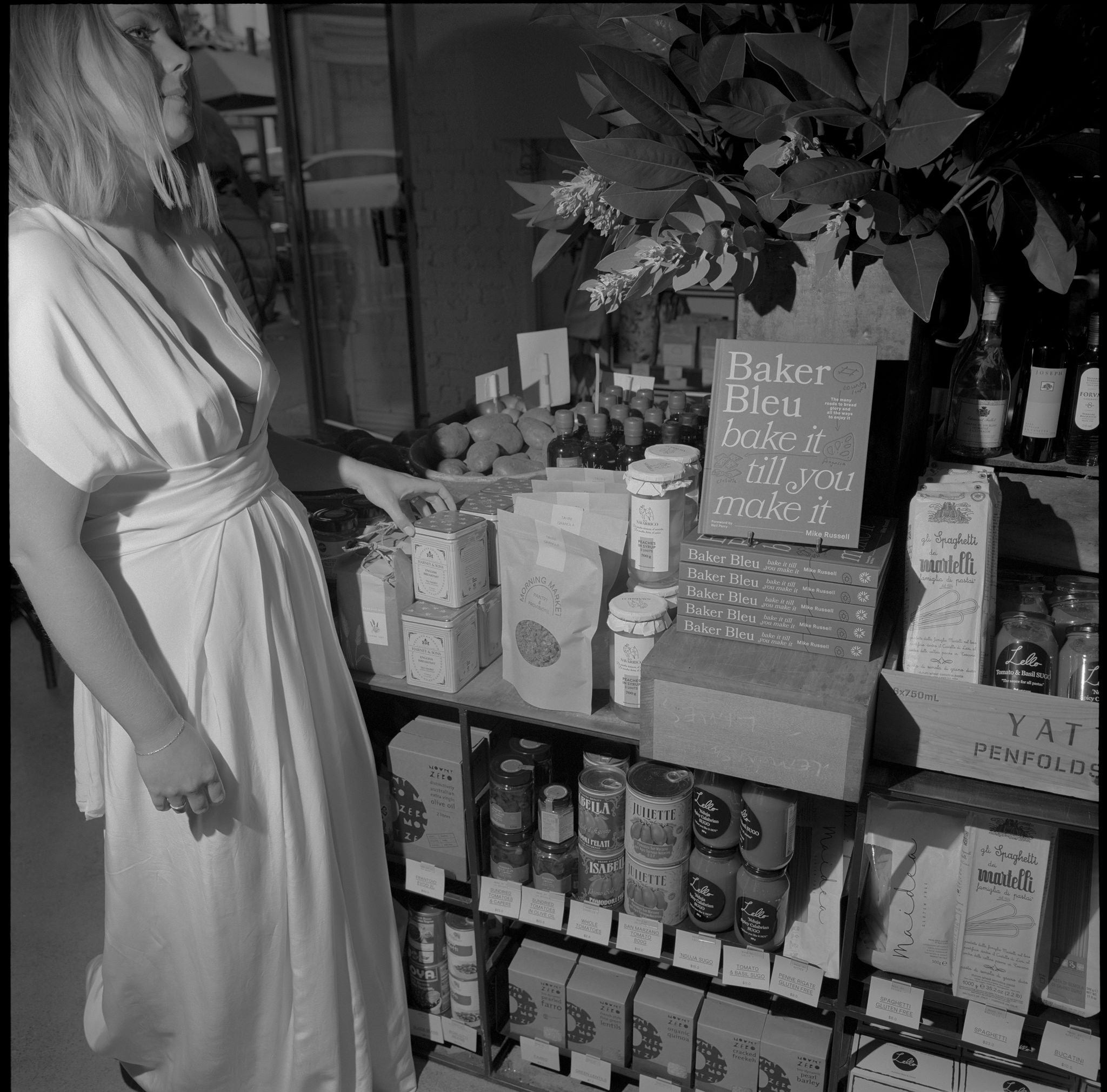 Bride in a minimalist white gown exploring a boutique market during her Recoleta wedding in Melbourne. The scene captures her browsing through shelves stocked with artisanal products, including gourmet foods, breads, and wine, under the soft light of a rustic setting. The presence of fresh produce, flowers, and a 'Baker Bleu' sign adds a charming, non-traditional element to this wedding day moment. This intimate and candid image showcases the personalized, relaxed nature of a Recoleta wedding, blending urban sophistication with unique and eclectic wedding details in one of Melbourne's most iconic venues.