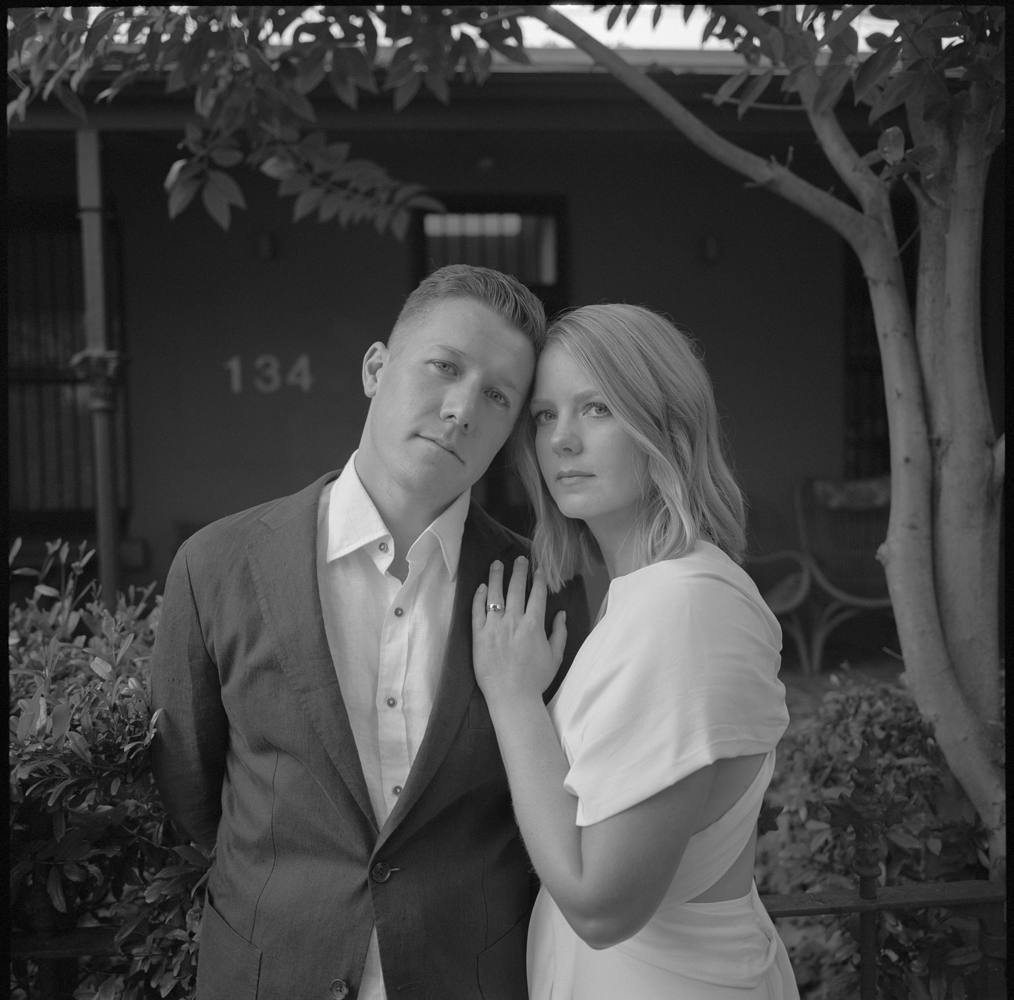 Close-up portrait of a bride and groom during their Recoleta wedding in Melbourne, standing outside a heritage home with the house number '134' visible in the background. The groom, dressed in a tailored suit, leans slightly into the bride, while the bride, wearing an elegant white gown, gently places her hand on his chest, showcasing her wedding ring. The intimate expression on their faces, combined with the greenery and historic setting, reflects the blend of timeless romance and modern simplicity that defines a Recoleta wedding. This photograph encapsulates the quiet elegance and personal connection that couples experience at their Recoleta Melbourne wedding.
