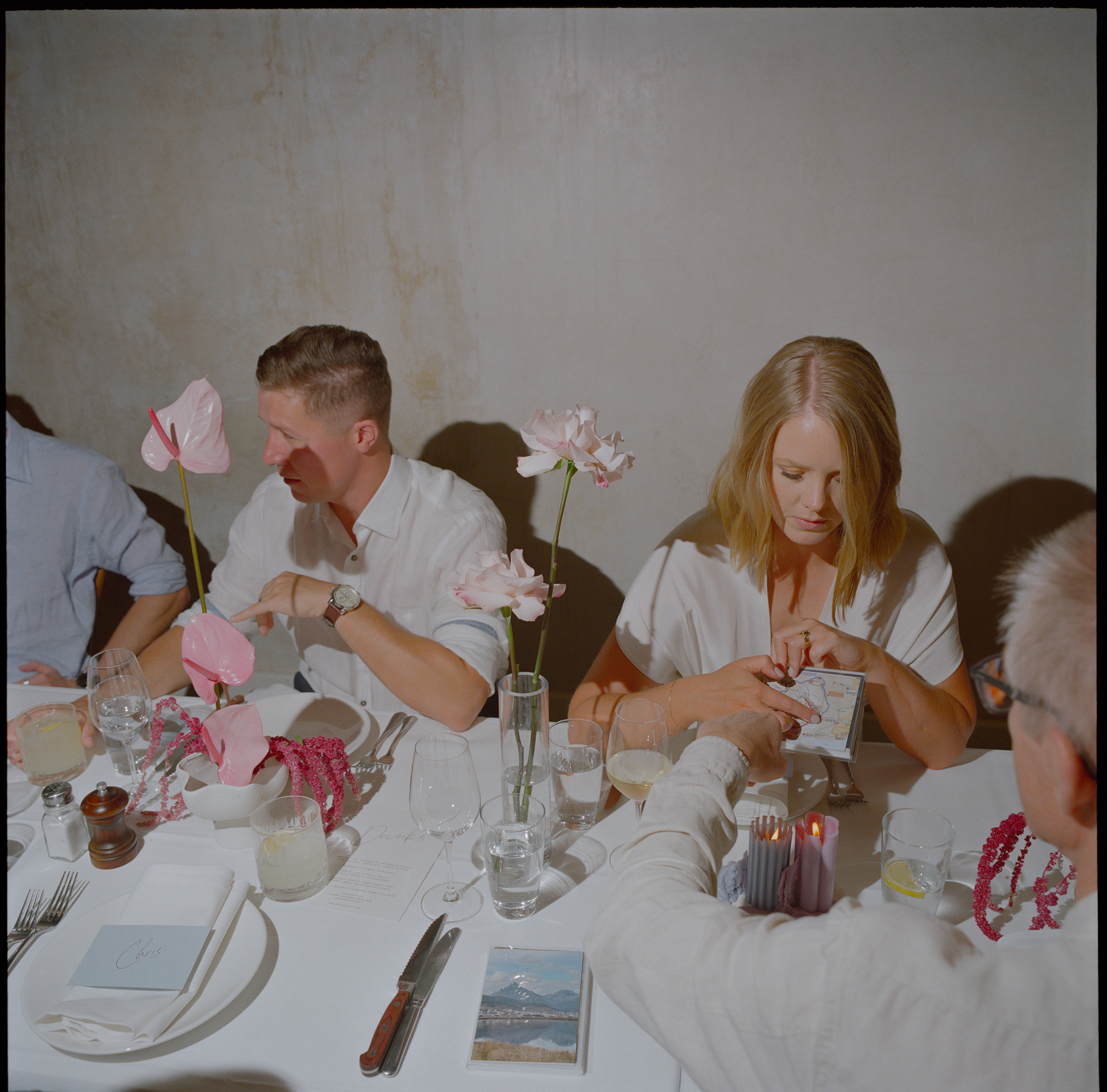 A lively and bright photograph capturing a group of guests seated around a table during a formal wedding at Recoleta in melbourne. The table is elegantly set with minimalist glass vases holding pink anthuriums and delicate roses, adding a touch of softness and color to the scene. One guest, a man in a white shirt, is engaged in conversation while a blonde woman next to him attentively examines a small booklet or card. The other guests around the table seem to be enjoying their drinks, with wine glasses, tumblers, and folded napkins neatly arranged on the white tablecloth. The soft lighting and muted background give the moment a relaxed yet refined atmosphere, encapsulating the essence of a joyful gathering.