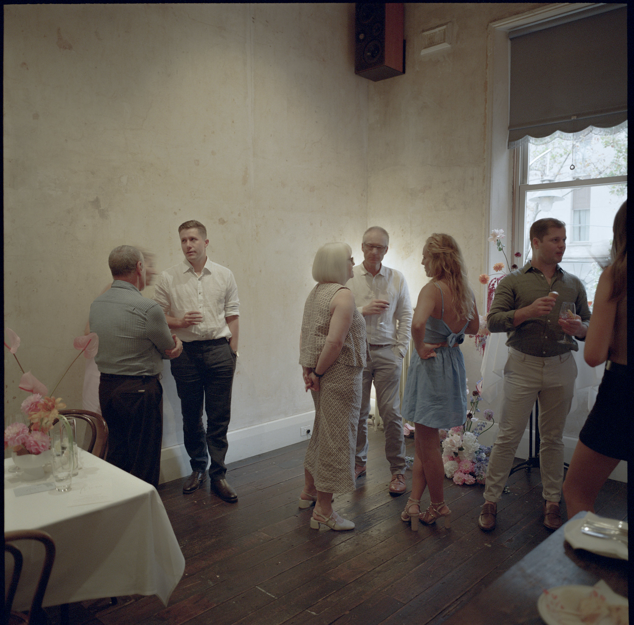 This photograph from a wedding at Recoleta in Melbourne captures a relaxed mingling moment between guests during the reception. Set against the backdrop of a rustic, aged wall with peeling paint, a small group of well-dressed guests engage in quiet conversation, drinks in hand. To the left, two men stand talking with an older woman who sports a chic bob haircut and wears a patterned, light-colored outfit. Next to them, a couple continues their conversation, both dressed casually yet stylishly. The man in the center wears a crisp white shirt, while the woman next to him dons a light blue dress with her back facing the camera, her long blonde hair flowing naturally.

The scene is softly lit, with daylight filtering in from a large window at the right side of the room. In front of the window stands a floral arrangement, a continuation of the colorful, understated floral theme that runs through the wedding, featuring pinks, lavenders, and oranges. The dark wooden floors and the muted tones of the walls lend a vintage charm to the space, while the simplicity of the decor allows the natural interactions between guests to take center stage. The overall ambiance is relaxed and intimate, with guests casually gathered in conversation, embodying the essence of a laid-back yet elegant wedding celebration.