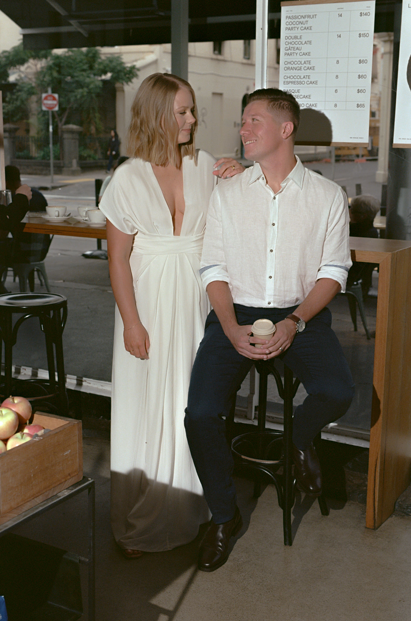 Bride and groom enjoying a quiet, candid moment during their Recoleta wedding in Melbourne, captured outside a charming local café. The bride, in a minimalist white gown, stands beside the groom, who is seated on a stool holding a cup of coffee, dressed casually in a white shirt and dark trousers. The couple shares a joyful glance, exuding relaxed happiness amidst the urban backdrop of Melbourne's city streets. The café menu and rustic decor, along with fresh apples displayed nearby, add to the modern, laid-back atmosphere of the scene. This image captures the essence of a Recoleta wedding—intimate, stylish, and effortlessly blending city life with romantic moments.