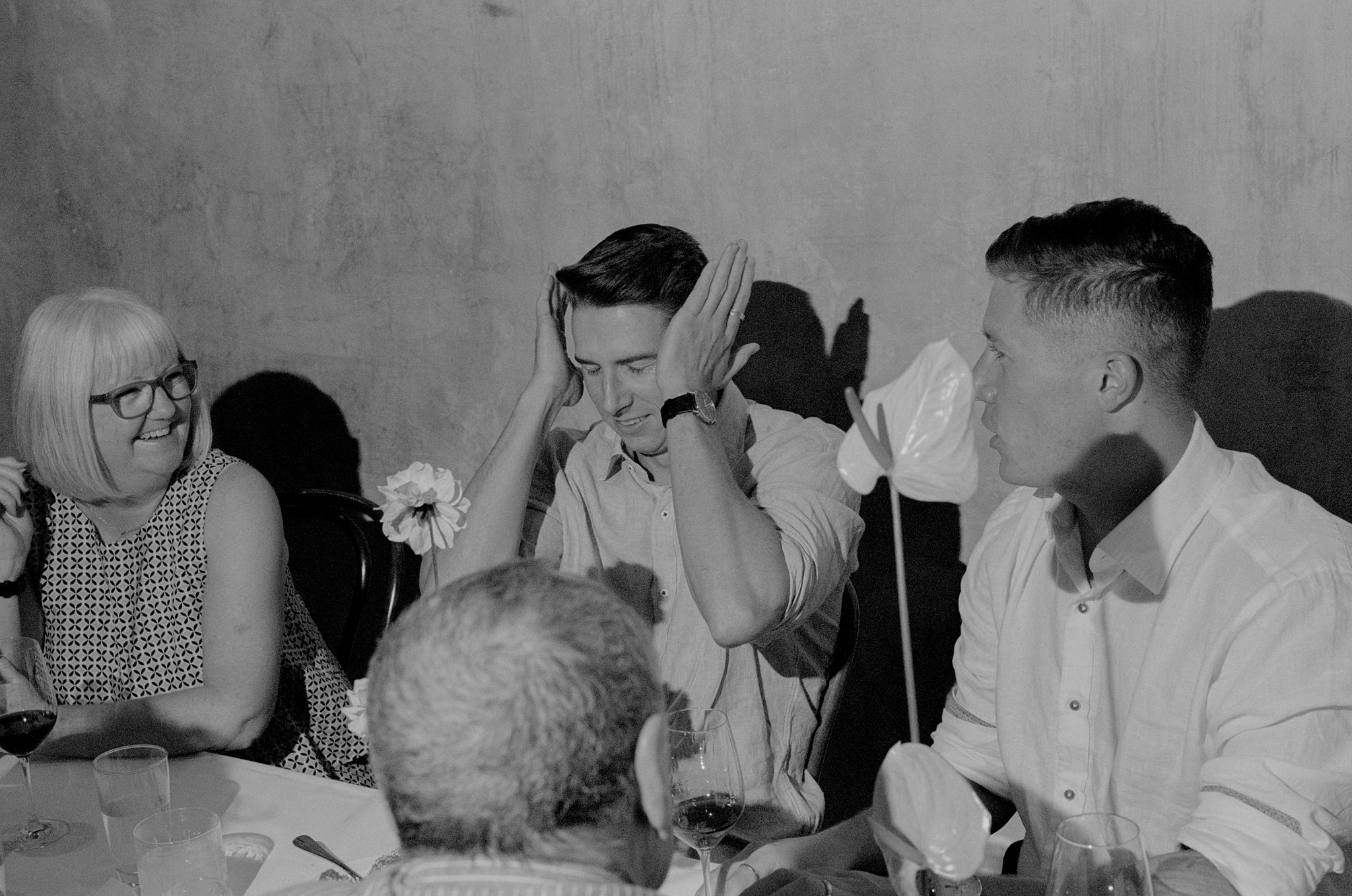 Guests at a wedding reception sharing a lighthearted moment, with one guest playfully covering his ears while smiling, seated between a woman with glasses and another guest in a white shirt. The table is adorned with elegant floral arrangements, including pink anthuriums, and glasses of wine. The casual and joyful atmosphere highlights the relaxed and celebratory vibe of the event, as guests engage in lively conversation and enjoy each other's company in a warm and intimate setting.